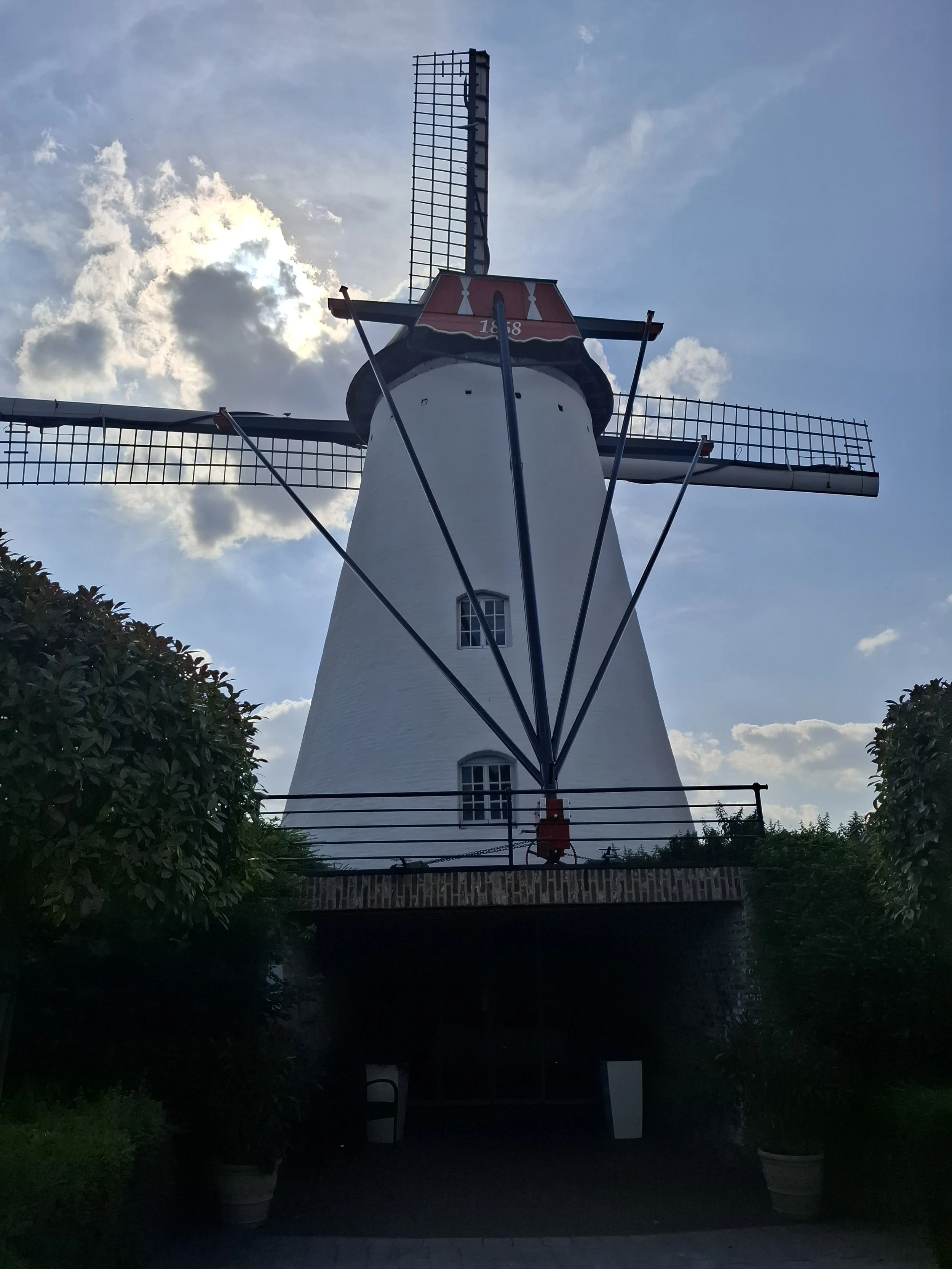 A white traditional windmill with four blades, a red and white top, and a small window near the bottom, set against a partly cloudy sky with the sun behind the clouds.