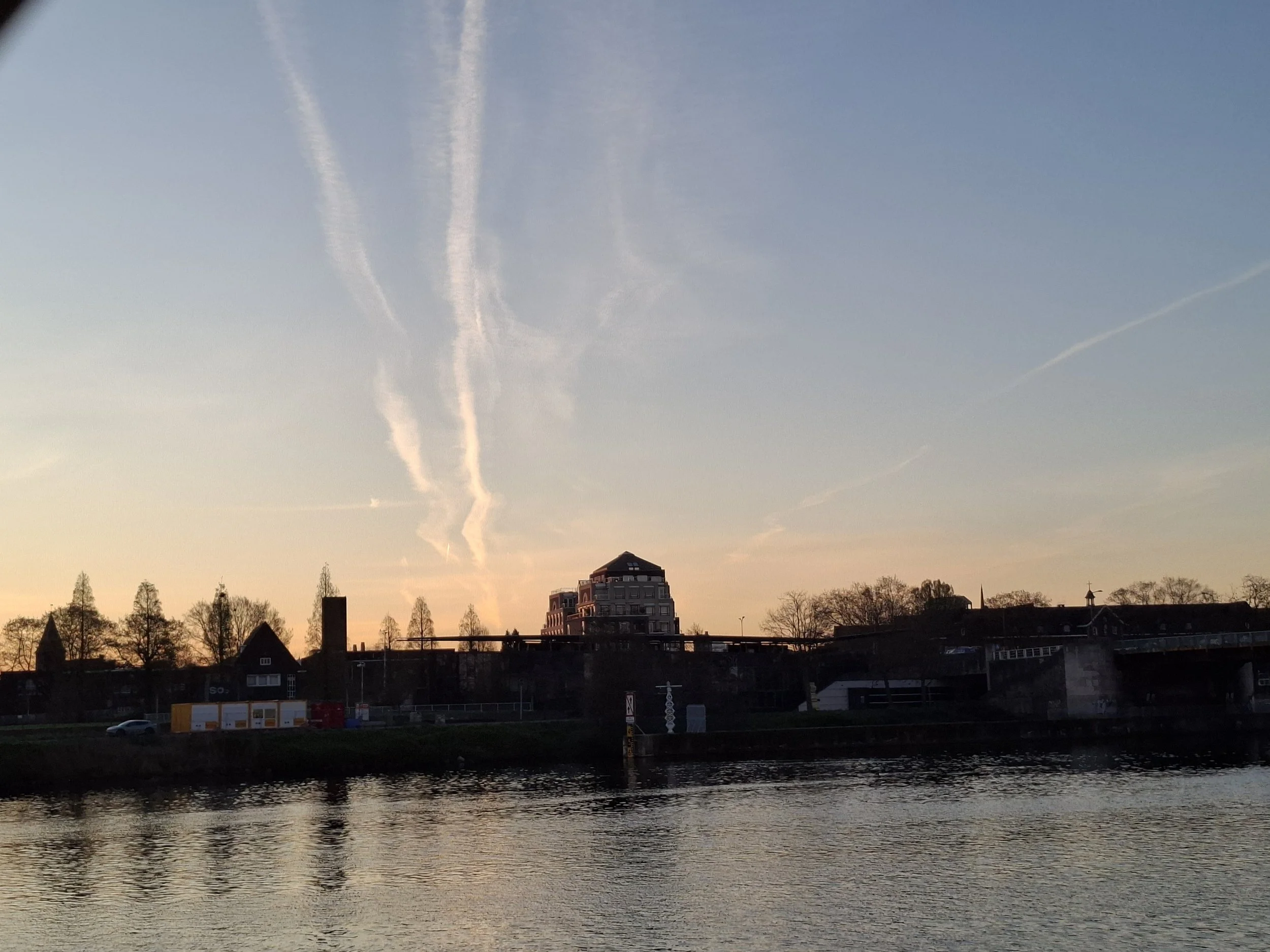 View of a river at sunset with silhouettes of buildings, trees, and a partly cloudy sky with contrails.