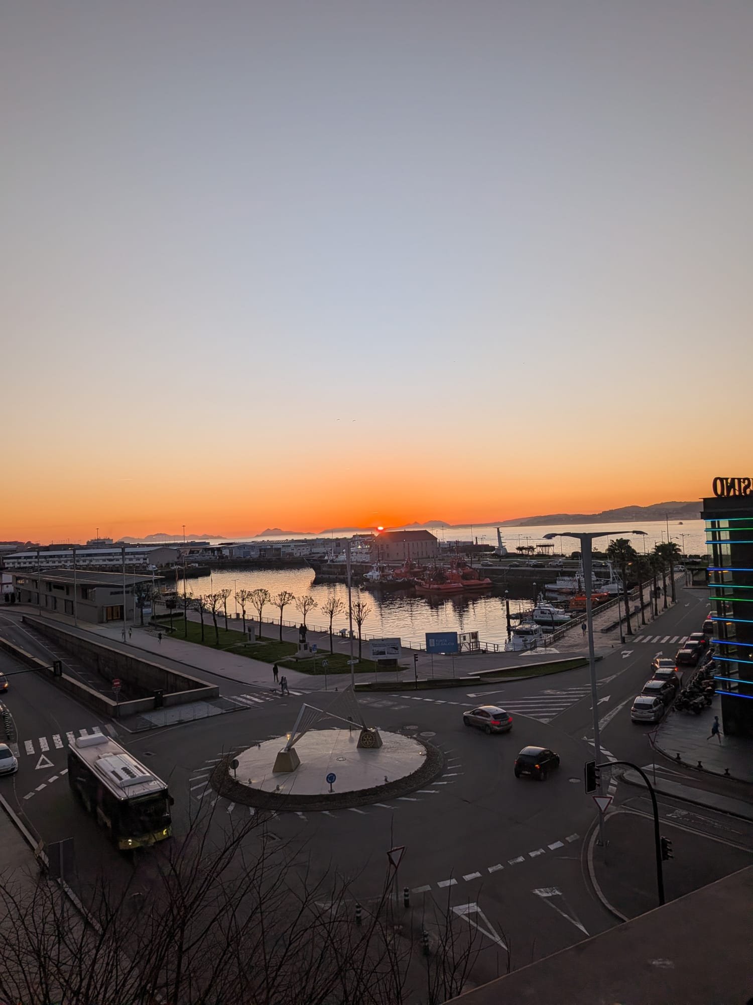 Sunset over marina with boats, water, and distant islands, viewed from a city street with cars and pedestrians.