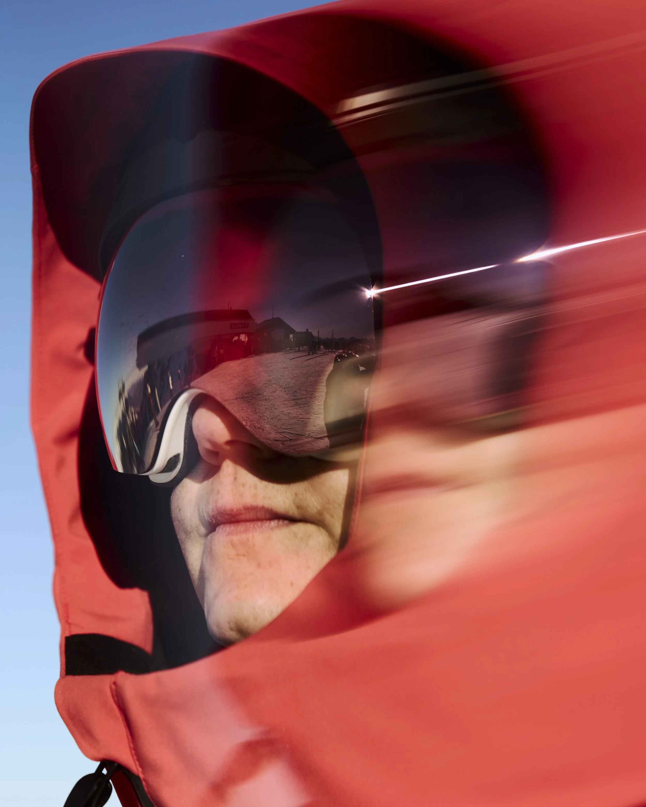 A close-up of an adaptive skier in black helmet and reflective ski goggles, with a red jacket, outdoors on a sunny day.