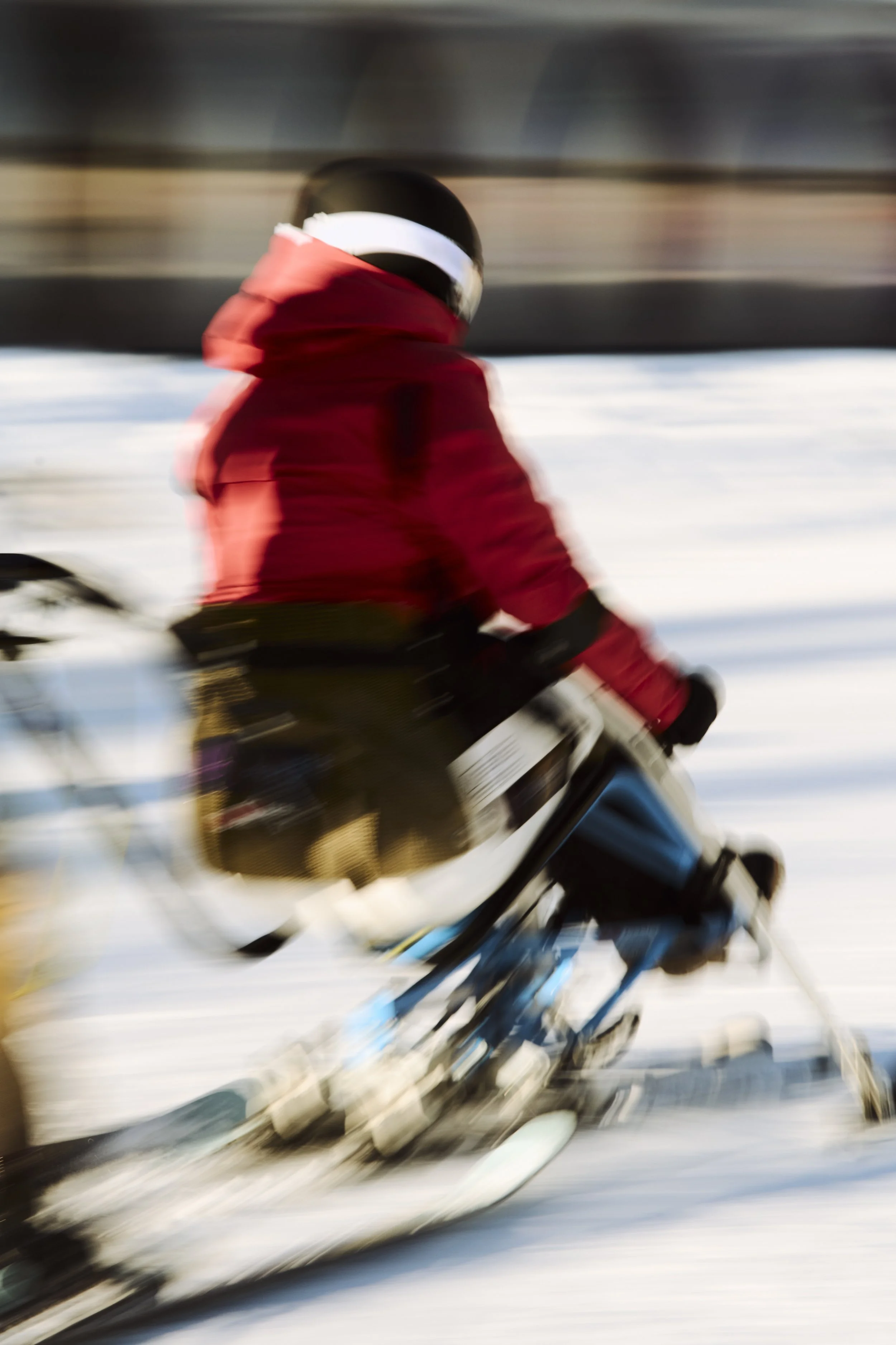 An adaptive skier rides down snowy terrain in a red jacket and black helmet with motion blur.