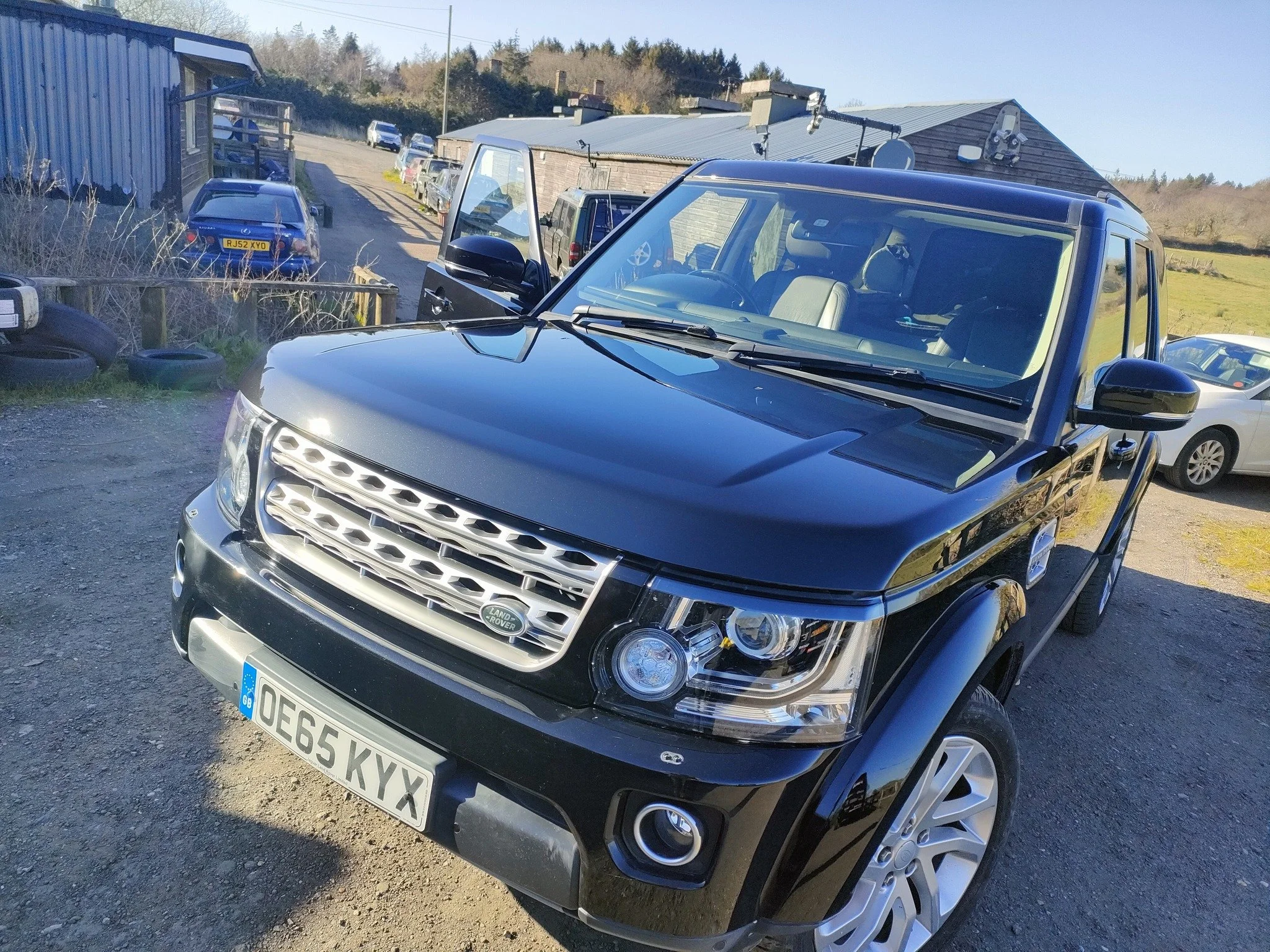 A black Land Rover SUV parked outdoors on a gravel surface with other cars and a blue building in the background under a clear sky.
