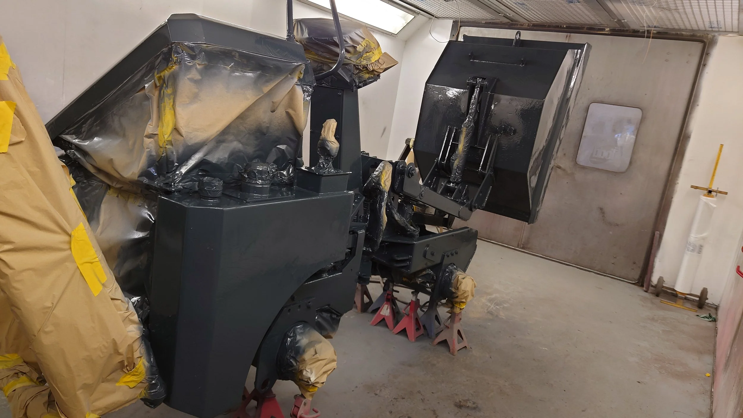 A machine in a workshop is masked with brown paper and tape, with several parts wrapped for painting or coating, supported by jack stands and set in a corner of the room.