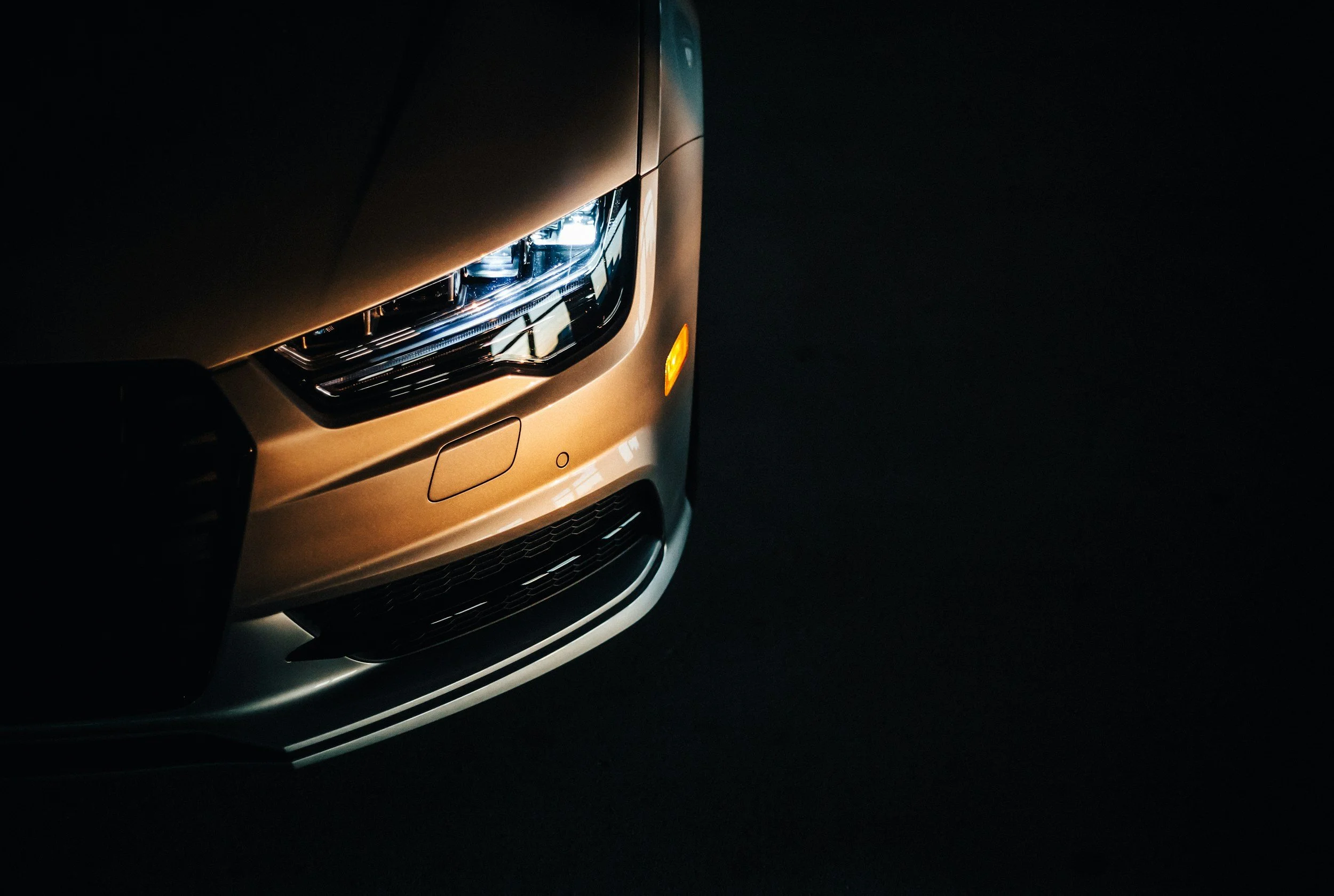 Close-up of the front left side of a luxury silver car, highlighting the headlight, grille, and bumper against a dark background.