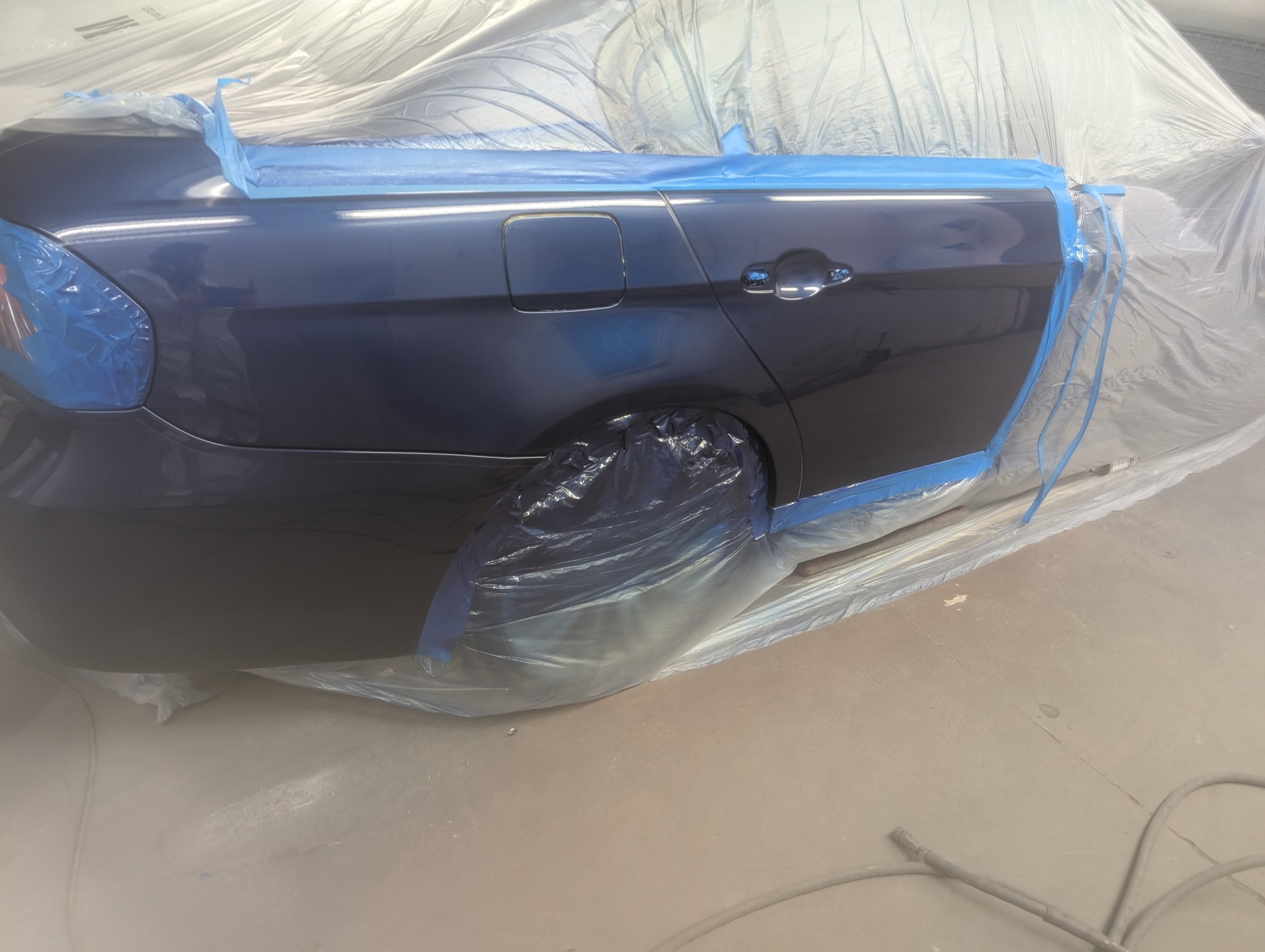 A dark-colored sedan car is covered and masked with plastic and painter's tape, likely undergoing painting or repair in an auto body shop.