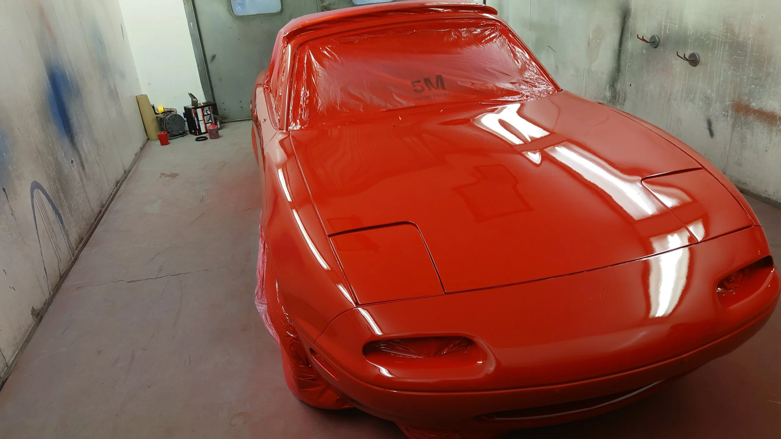 Red sports car inside a garage, covered with red protective wrap on the sides, with the windshield and windows uncovered.