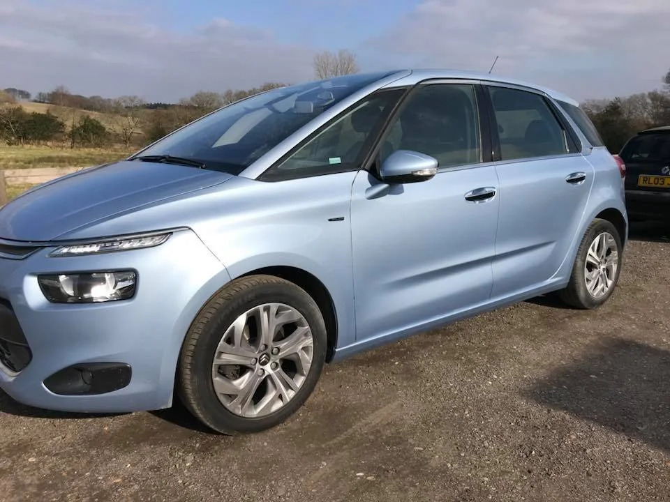 A light blue hatchback car parked on a gravel lot with a rural landscape and cloudy sky in the background.