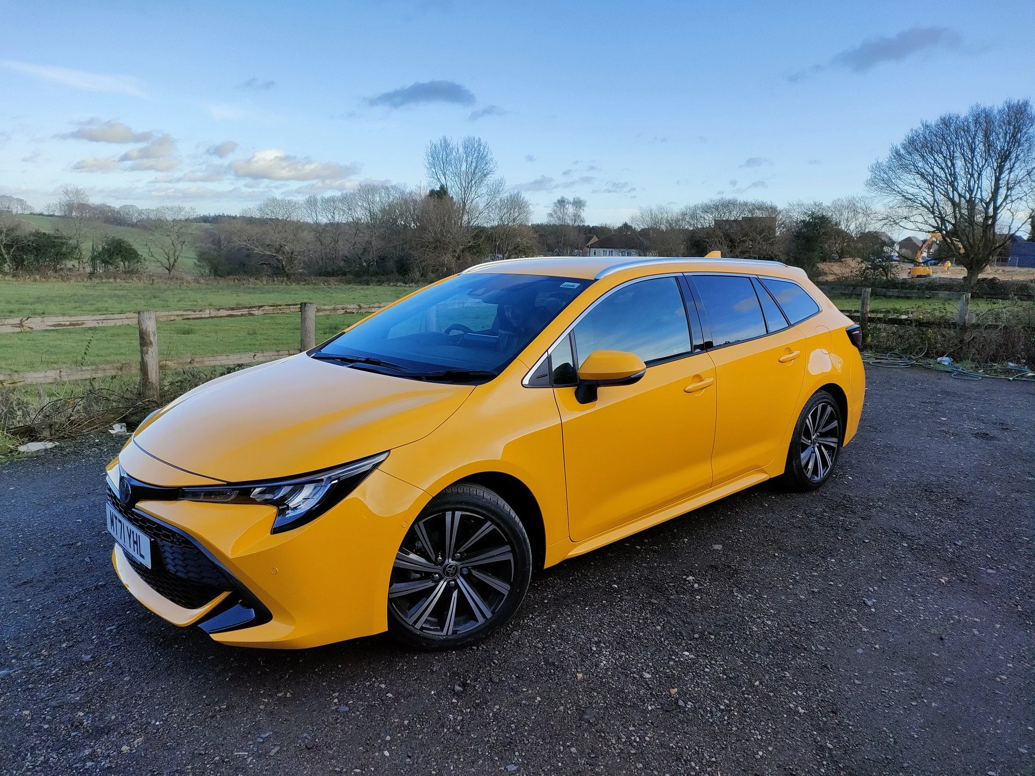 Yellow hatchback car parked on a gravel surface with a rural landscape, trees, and houses in the background under a partly cloudy sky.