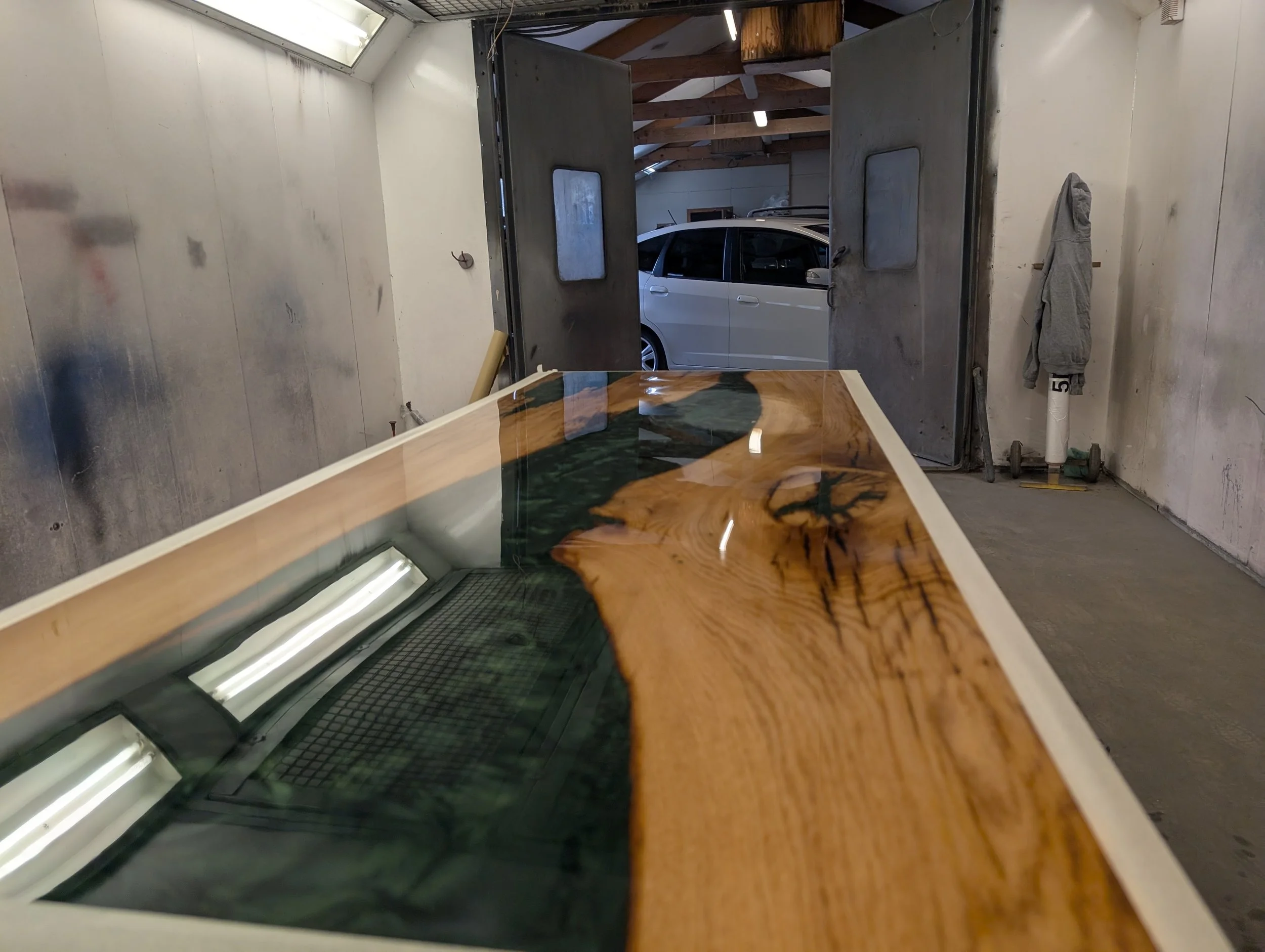 A custom-made wooden and glass table inside a workshop with a white vehicle visible through the open door in the background.