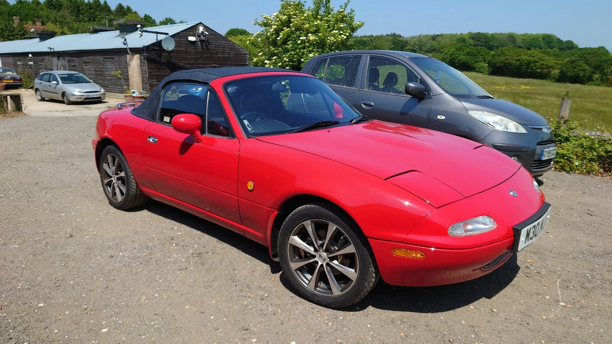 Red Mazda MX-5 Miata convertible parked on gravel with a black hardtop, alongside a gray compact car, in a rural setting with a wooden building and green hillside in the background.