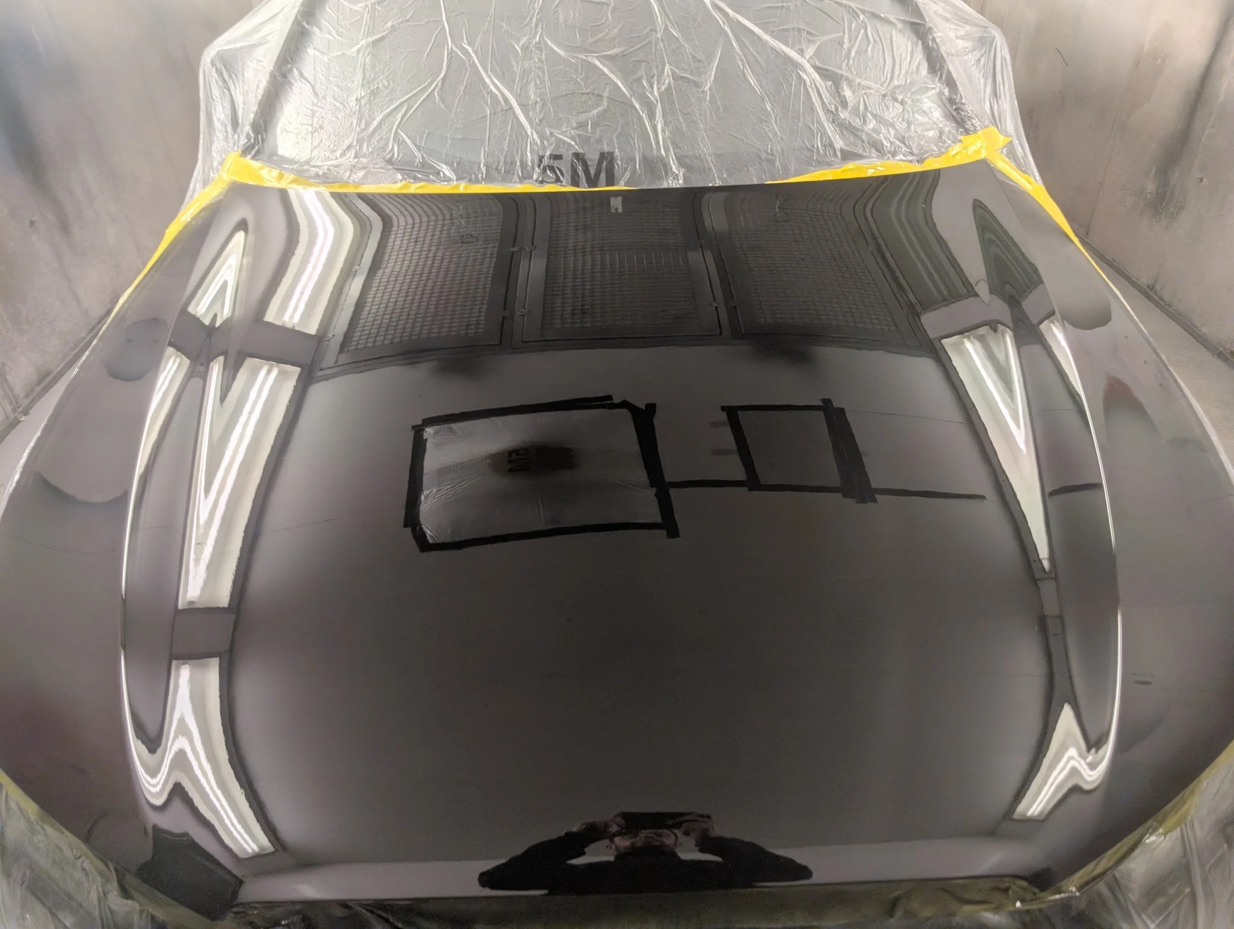 A reflective, highly polished, metallic car hood with black tape markings on it, surrounded by masking and painter's tape in a workshop.