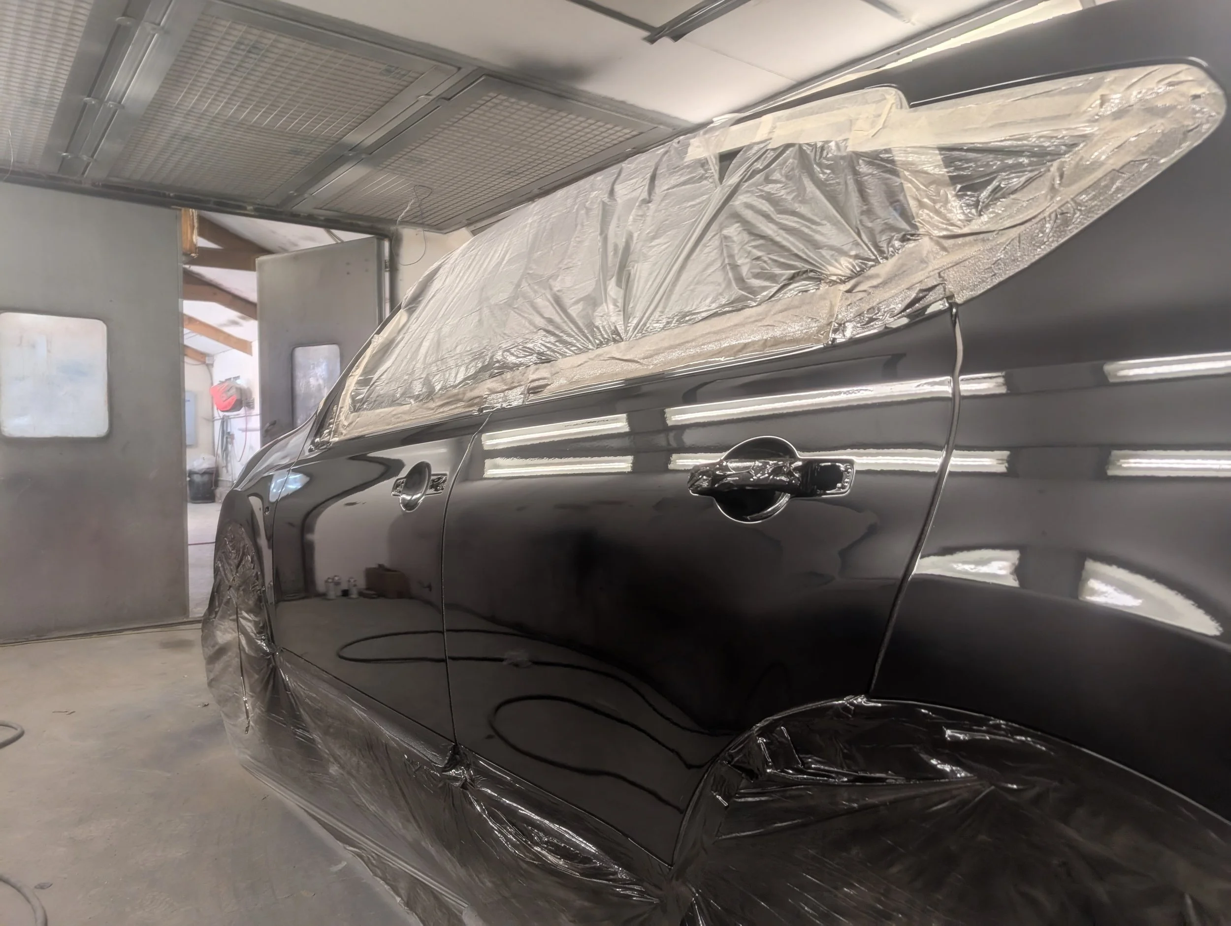 A black car inside a spray booth, covered with protective plastic sheeting and masking tape for painting.