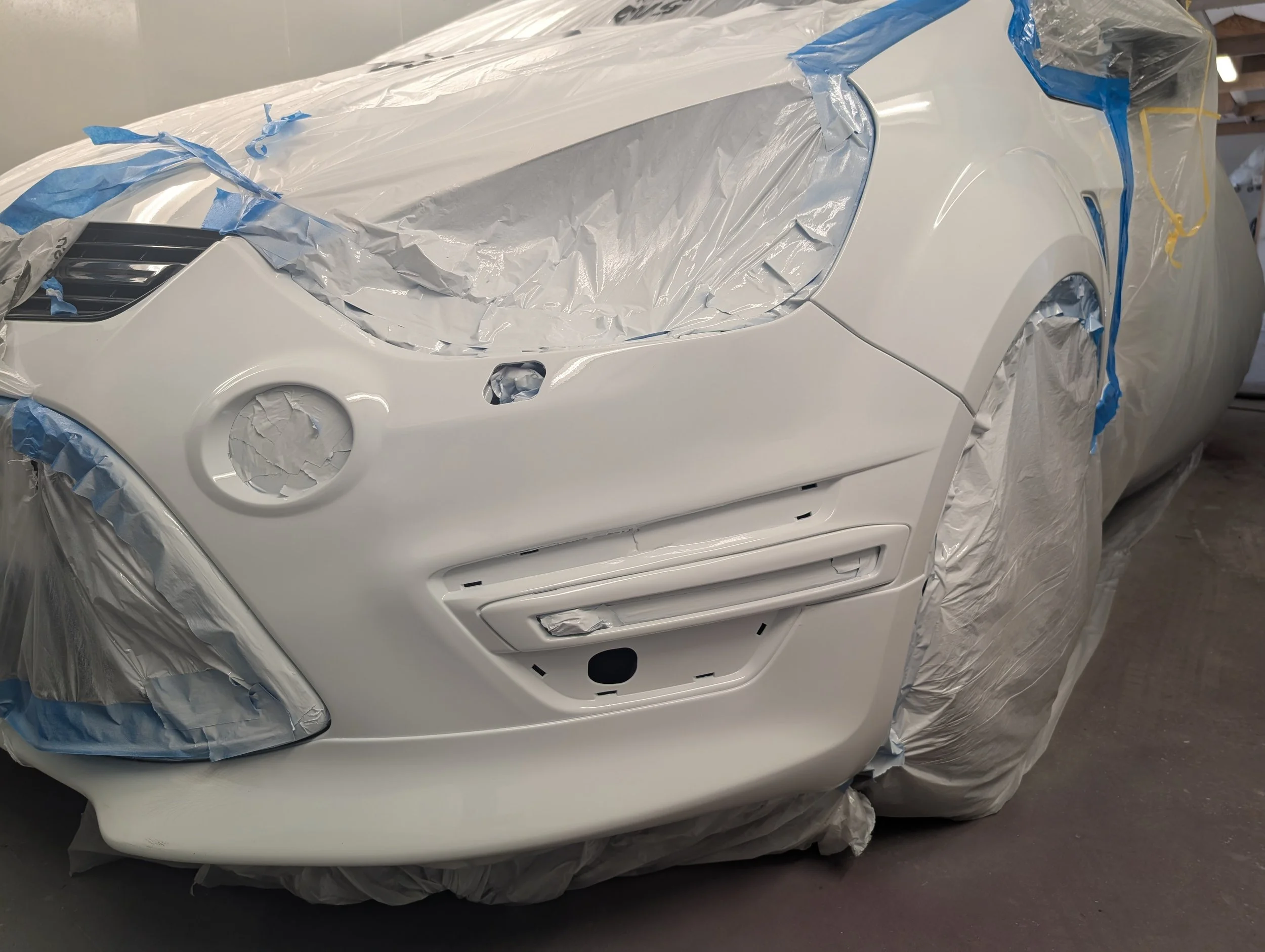 White car undergoing painting, with door and headlight areas masked off with plastic and tape in a workshop.