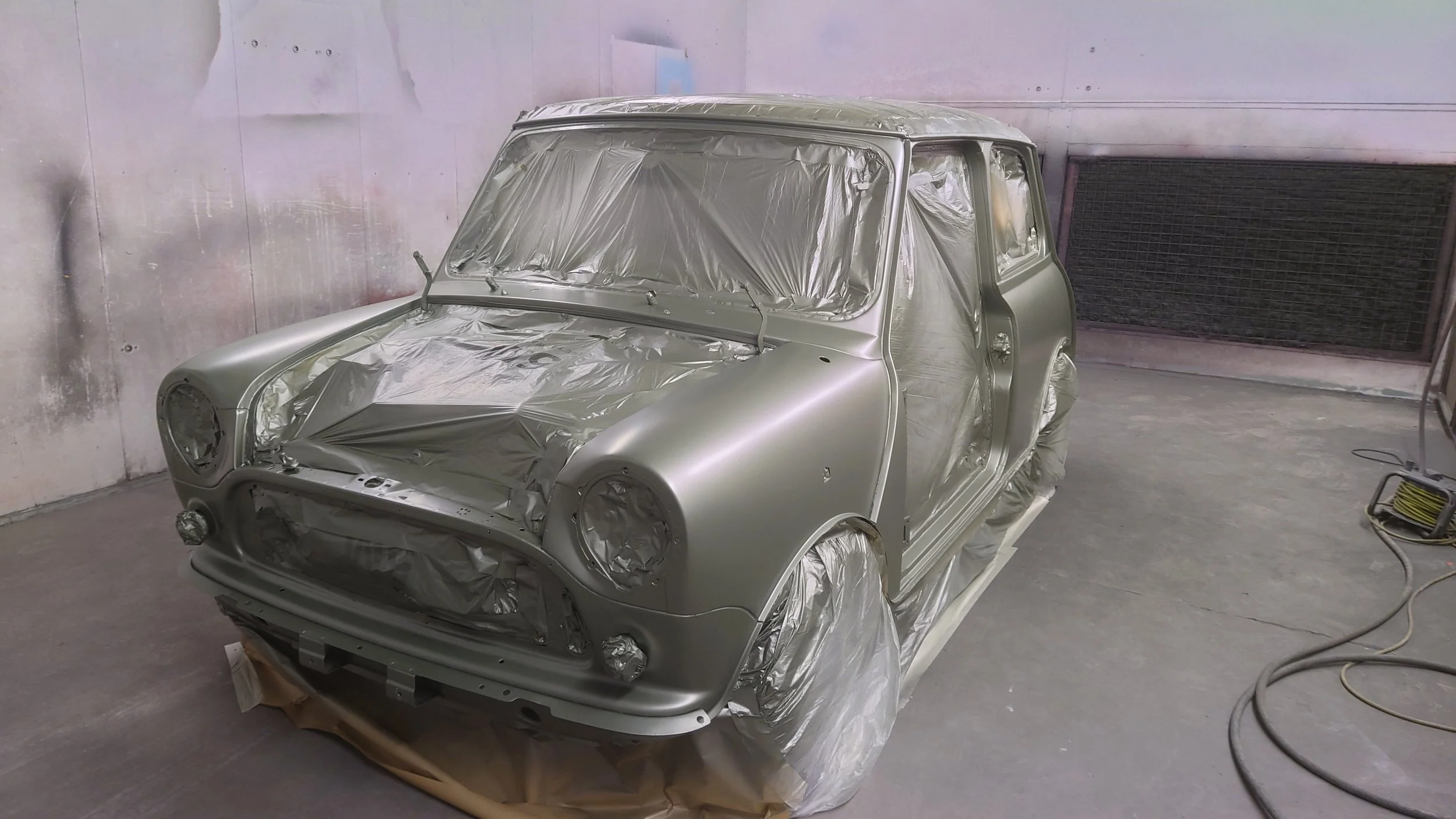 A mini car in a paint booth, coated in primer with masking paper and foil covering its windows, headlights, and wheels during a painting process.
