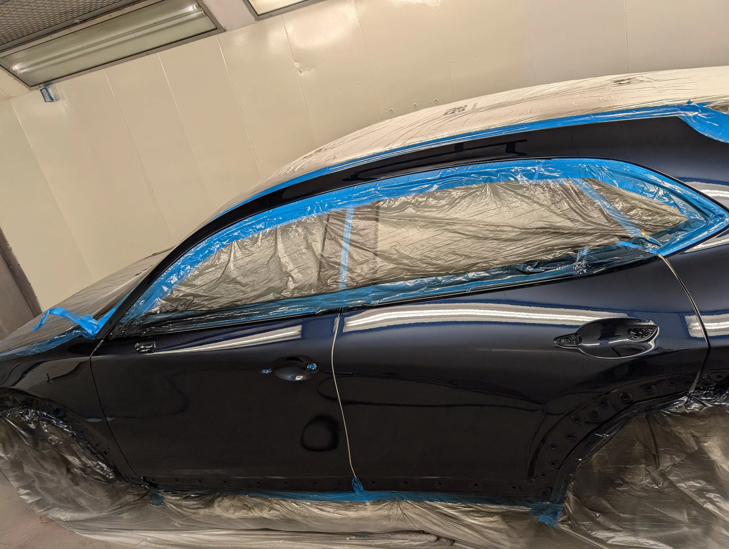 A black car in a paint booth with paper and blue painter's tape covering the windows and surrounding areas.