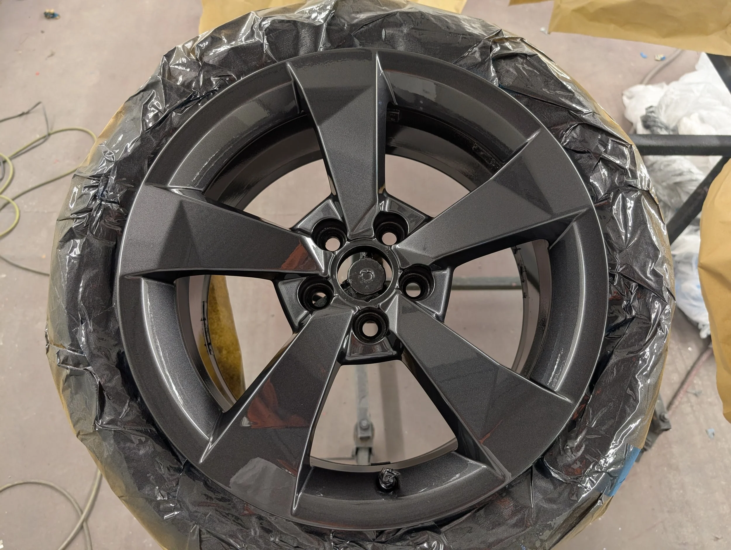 A car wheel with a dark metallic finish, wrapped in protective plastic, on a worktable in a workshop.