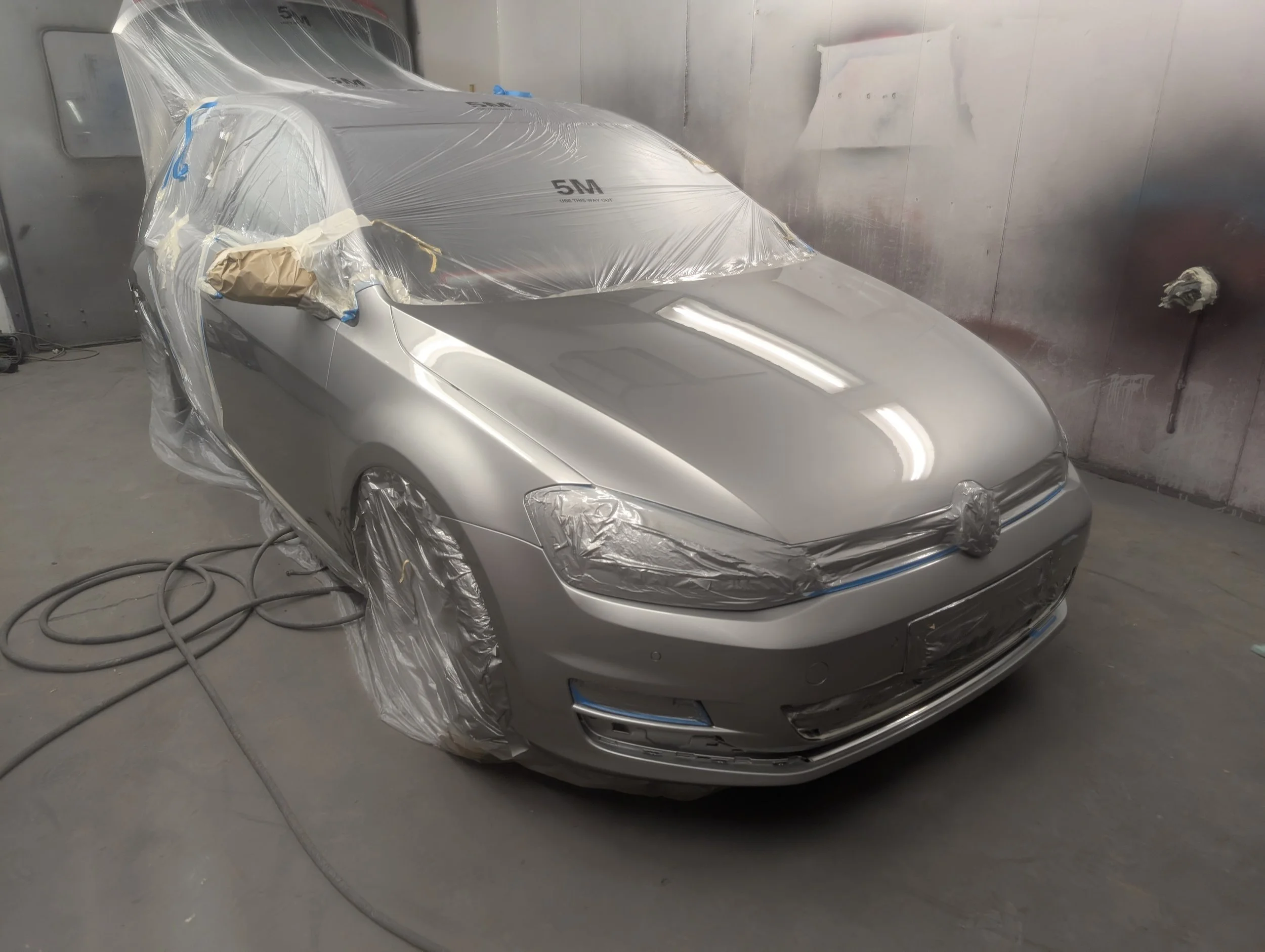 A silver car inside a paint booth, wrapped in plastic for a paint job, with masking tape covering certain areas, and a spray gun on the floor.