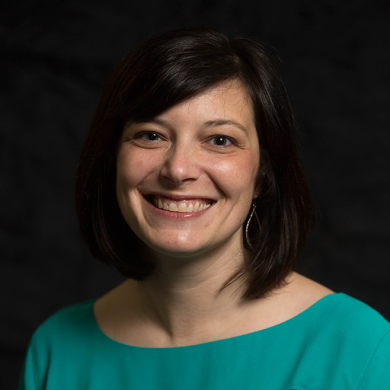 A woman with short dark brown hair smiling, wearing a teal top and hoop earrings, against a dark background.