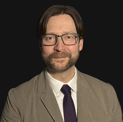 A man with glasses, a beard, and shoulder-length brown hair, wearing a beige blazer, white shirt, and dark tie, posed against a black background.