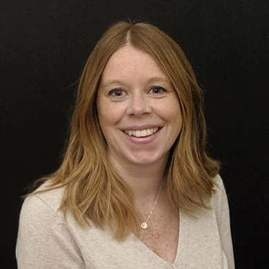 A woman with shoulder-length red hair smiling against a black background.