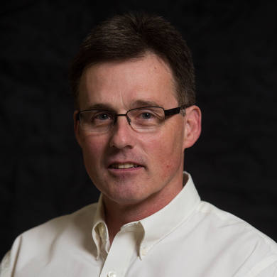 Headshot of a man with glasses wearing a white collared shirt against a dark background.