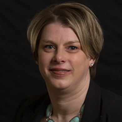 A woman with short Blonde hair wearing a black blazer and a turquoise necklace against a dark background.