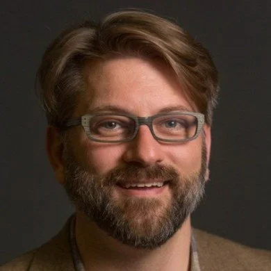 A man with glasses, brown hair, and a beard, smiling against a dark background.