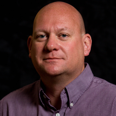Portrait of a bald man wearing a purple button-up shirt against a black background.