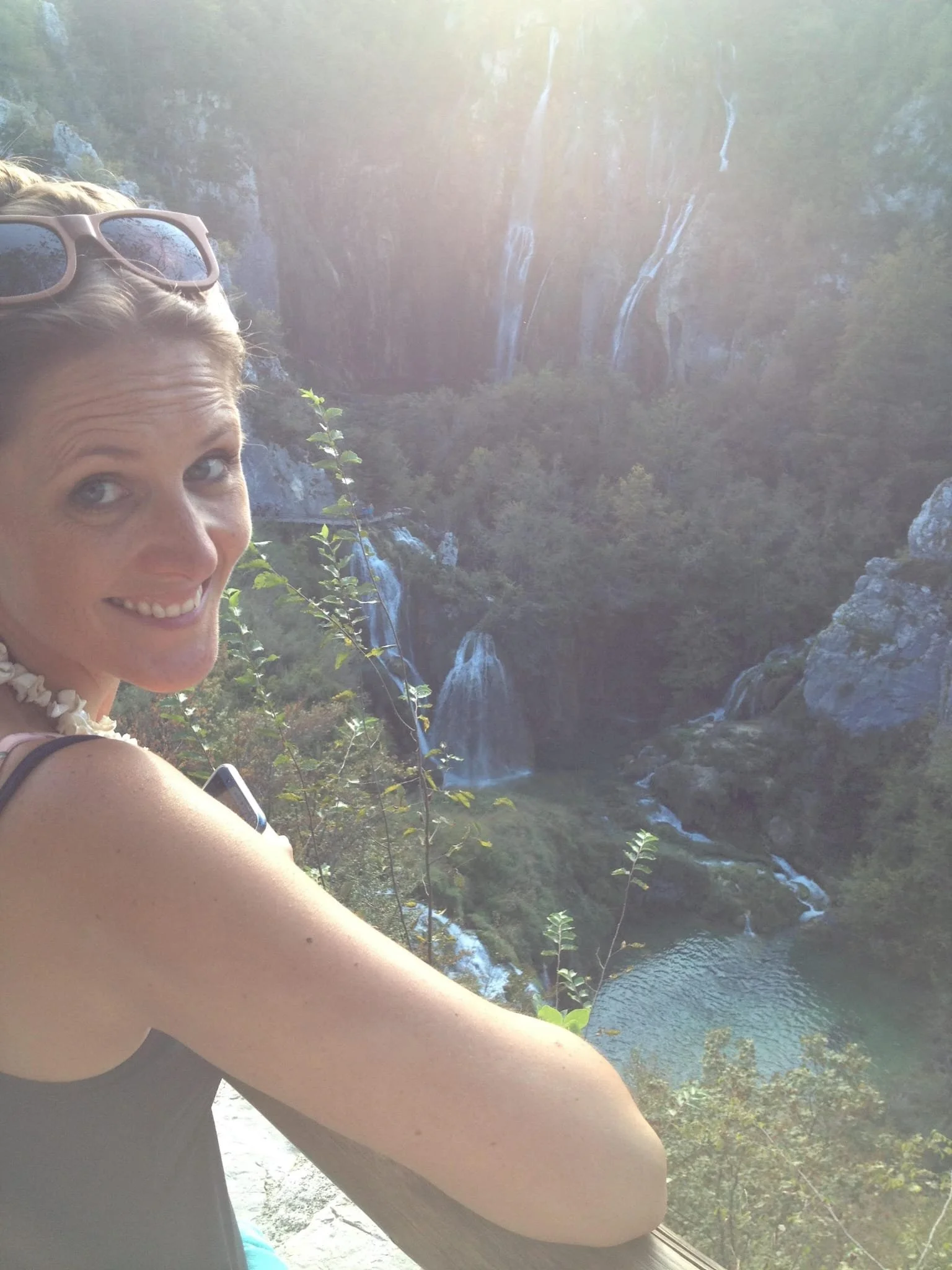 A woman with sunglasses on her head and a shell necklace smiling at the camera with waterfalls and a river in a forested canyon in the background.