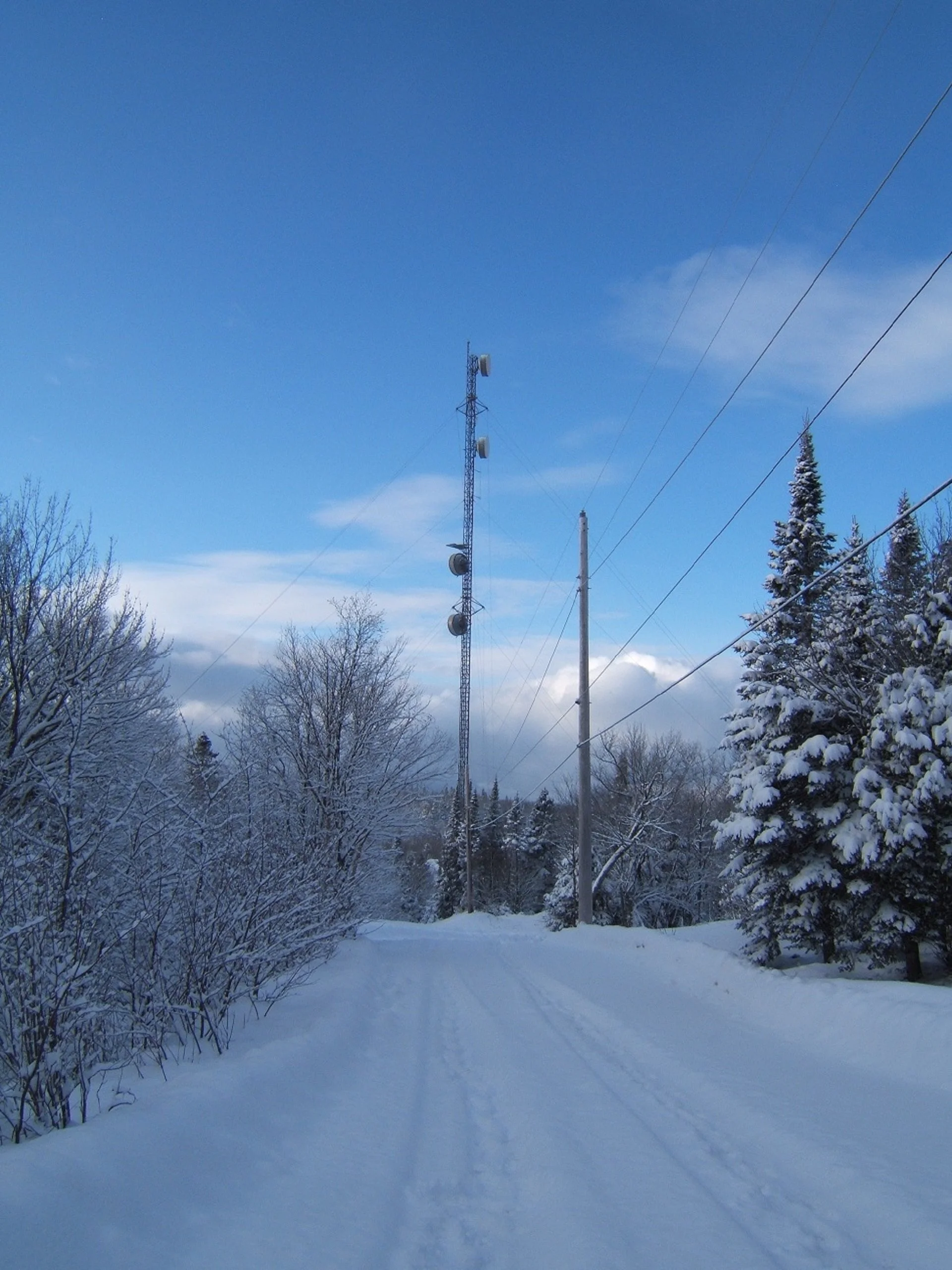 Snow-covered rural road with trees on both sides, power lines and a tall communication tower under a partly cloudy blue sky.