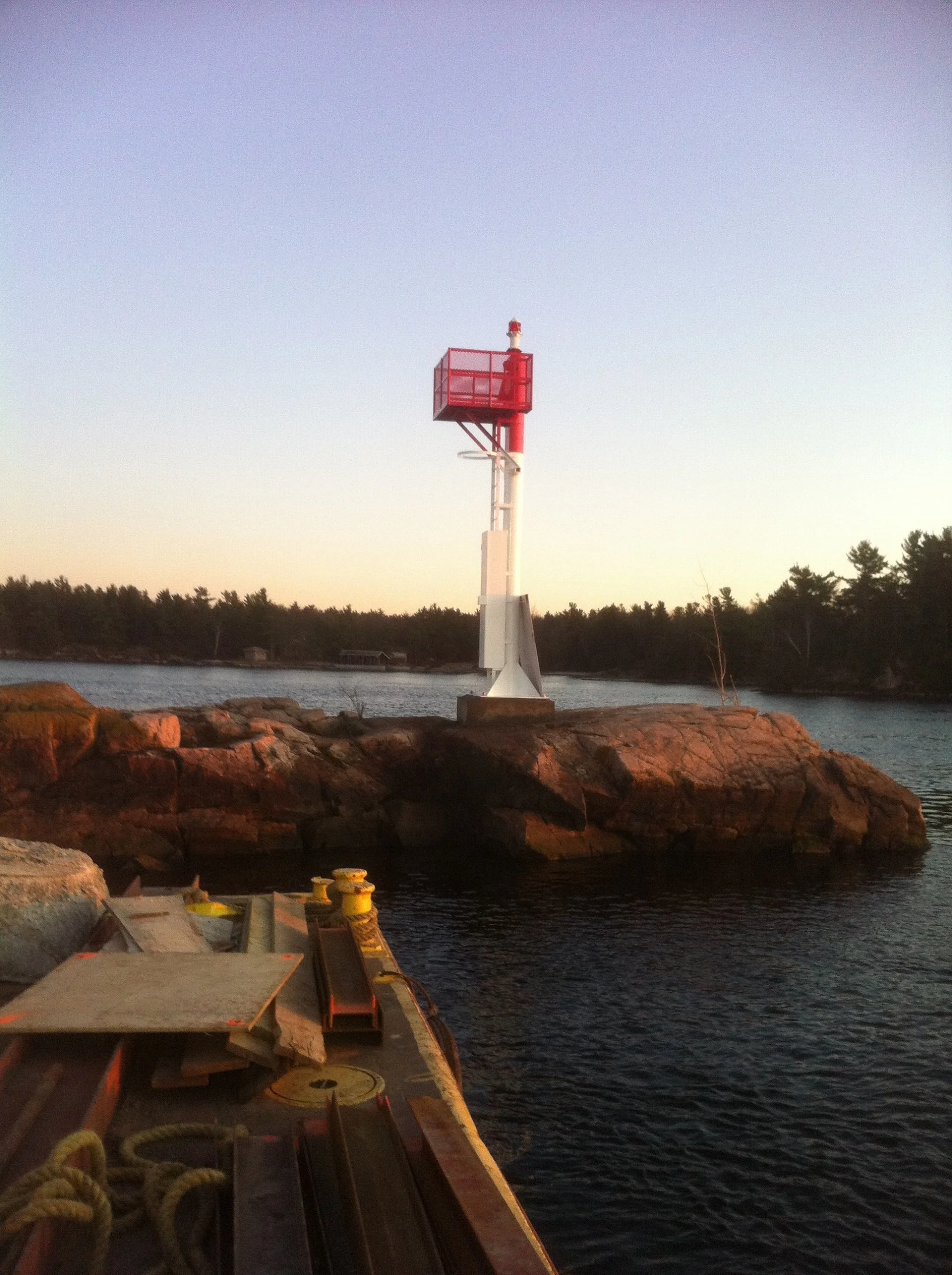 A short red and white communications tower on rocks near water with tools and supplies in the foreground, and trees in the background during sunset or sunrise.