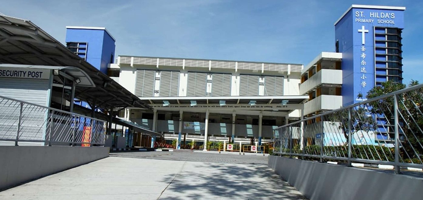 Exterior view of St. Hilda's Primary School building with blue accents and a cross symbol on the sign, including a security post and entrance driveway.