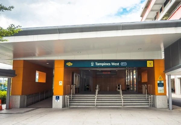 Entrance to Tampines West MRT station with stairs and escalator, orange and black exterior, and signage indicating station name and accessibility features.