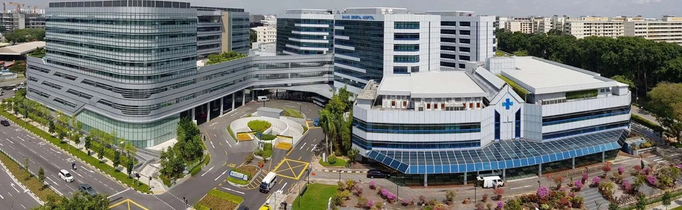 Aerial view of a modern hospital complex with multiple buildings, parking lots, and landscaped gardens.
