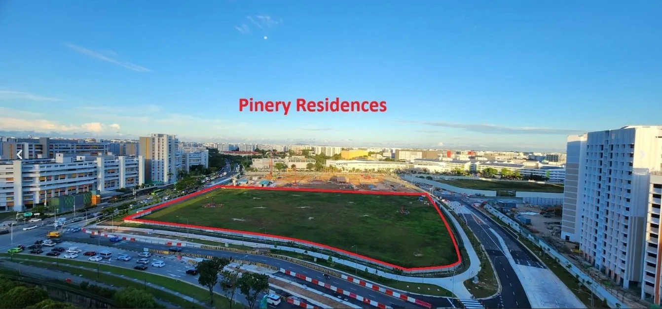 Aerial view of an urban area with a large empty green lot marked as Pinery Residences, surrounded by roads, modern buildings, and construction activity in the background under a blue sky.