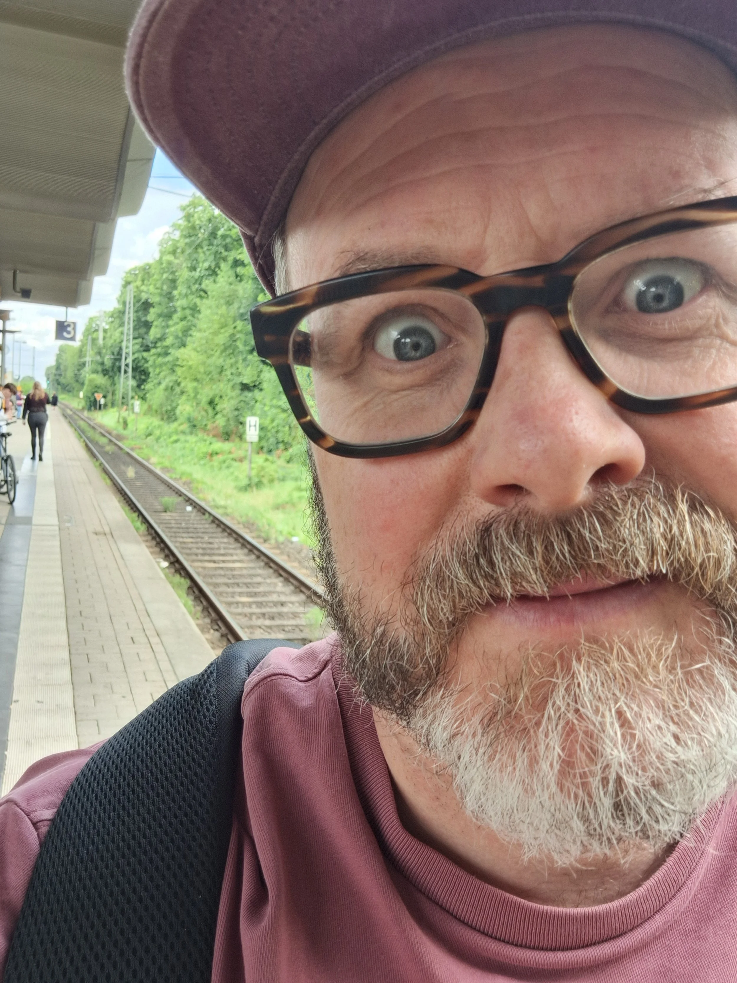 Close-up of a man with glasses, a beard, and a purple cap at a train station platform, with train tracks and other people in the background.