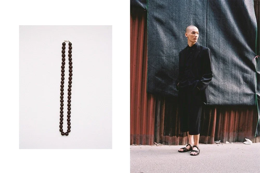A long black bead necklace on a white background, and a Vietnamese person with a shaved head wearing a black suit and sandals leaning against a weathered wall.