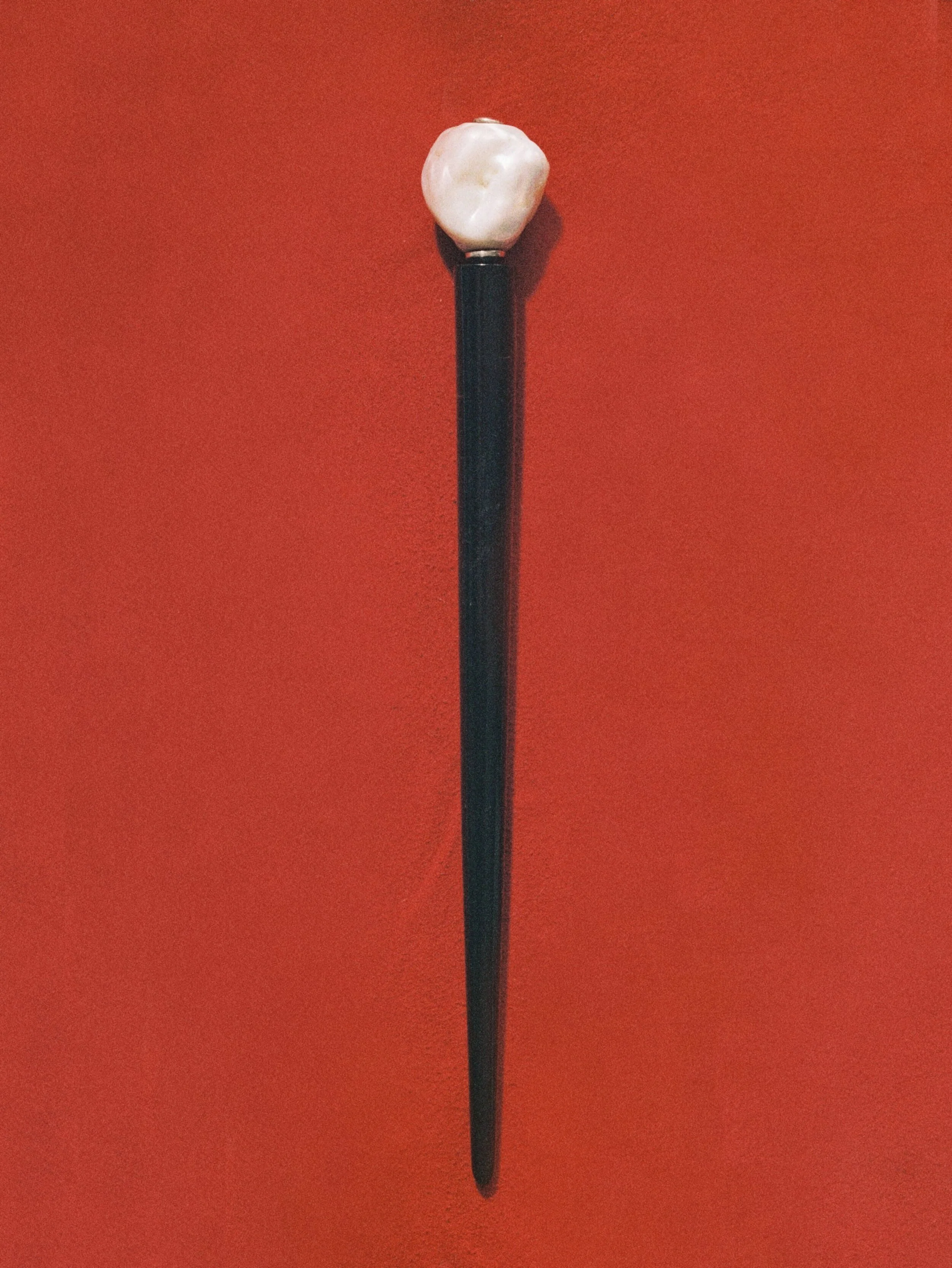 A black hairpin with a baroque pearl on the top against a red background.