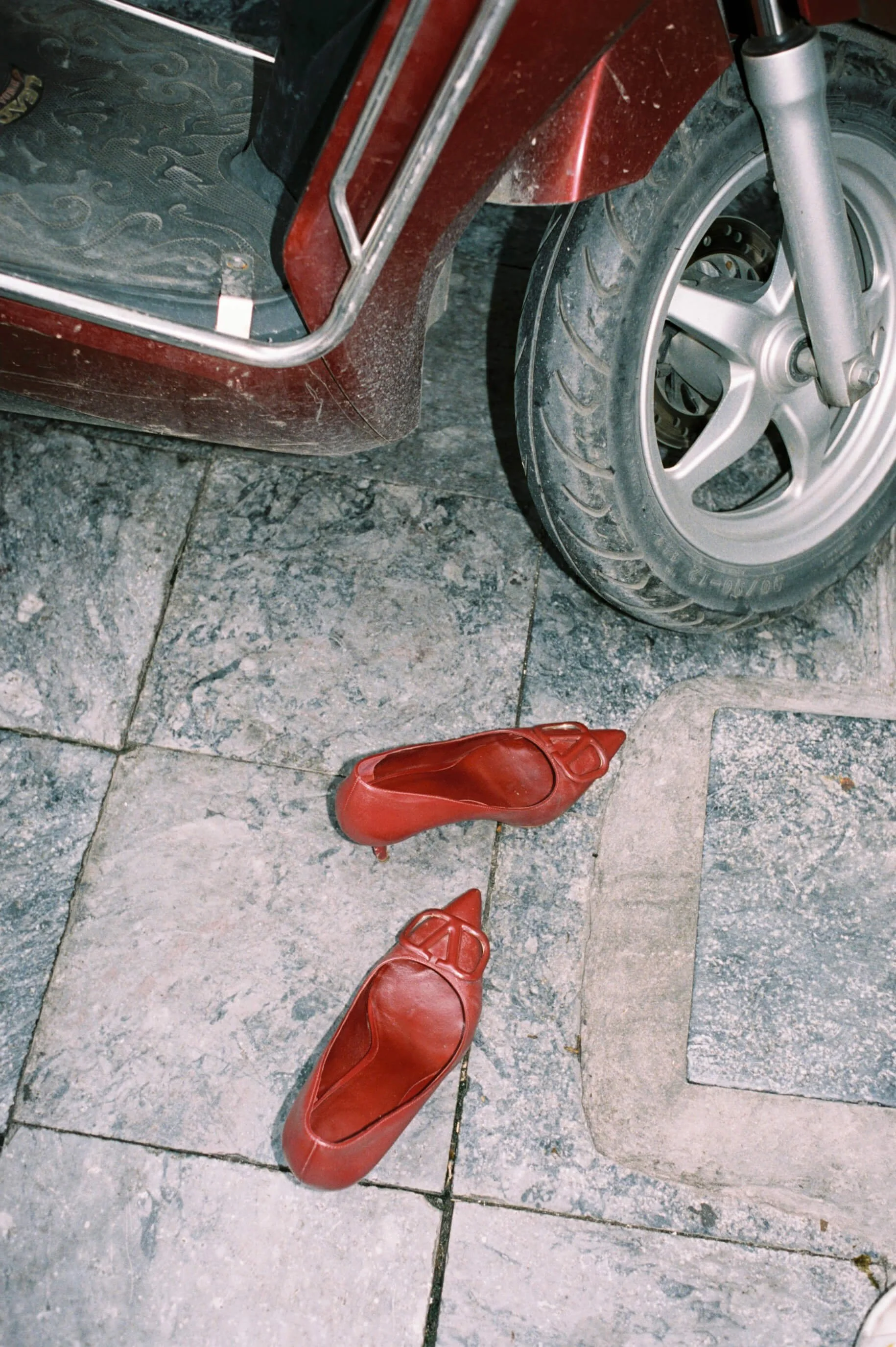 Red high-heeled shoes on a stone floor with a motorcycle tire nearby.