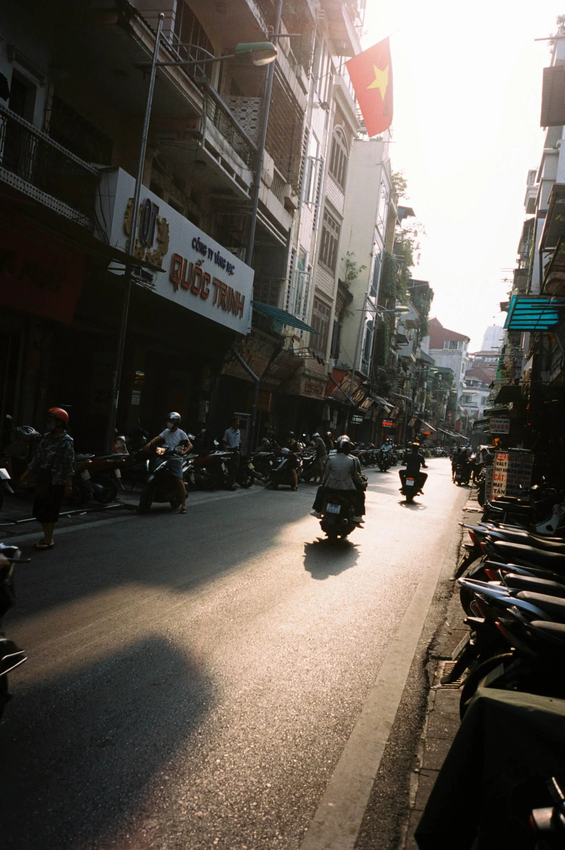 Street scene in an Asian city with buildings on both sides, motorbikes and scooters on the road, and the Vietnamese flag hanging from a building, with the sun setting or rising casting a warm glow.
