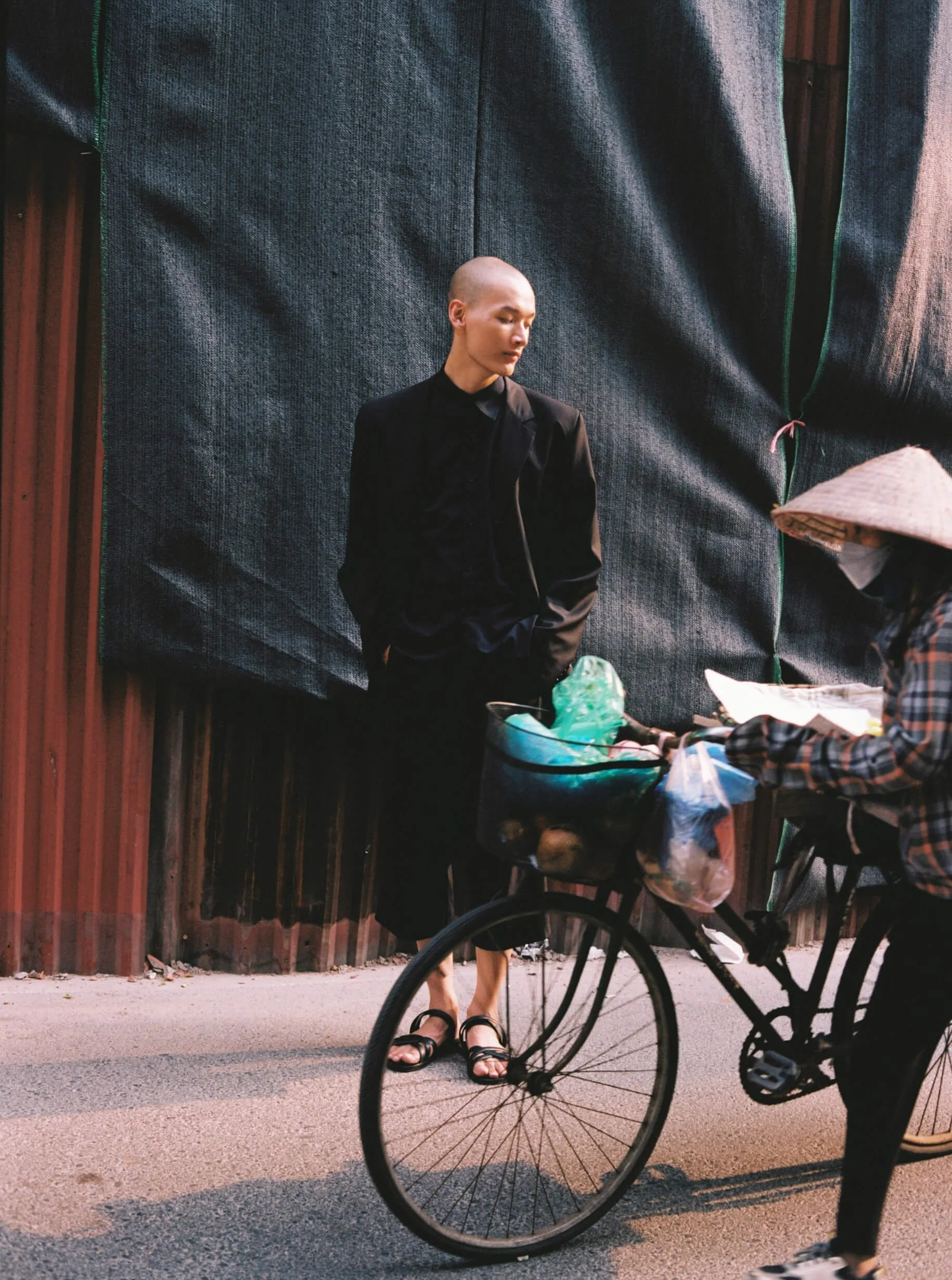 A person wearing a black suit and sandals standing against a large fabric backdrop, with a person on a bicycle partially visible in the foreground and a woman in a traditional conical hat and face mask looking at a table.