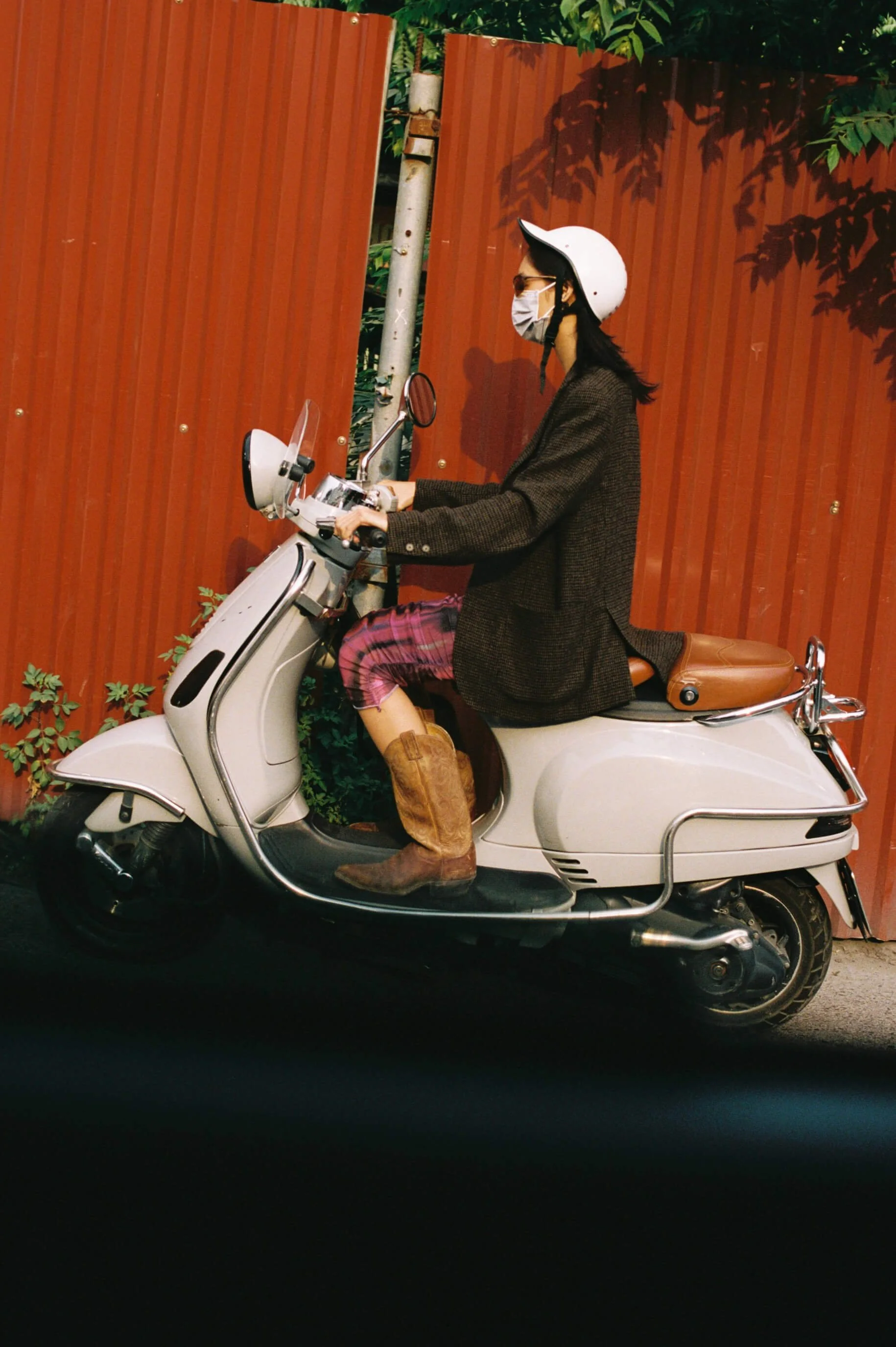 A person wearing a white helmet, face mask, sunglasses, and a dark coat riding a white scooter with a brown seat in front of a red metal fence and green foliage.