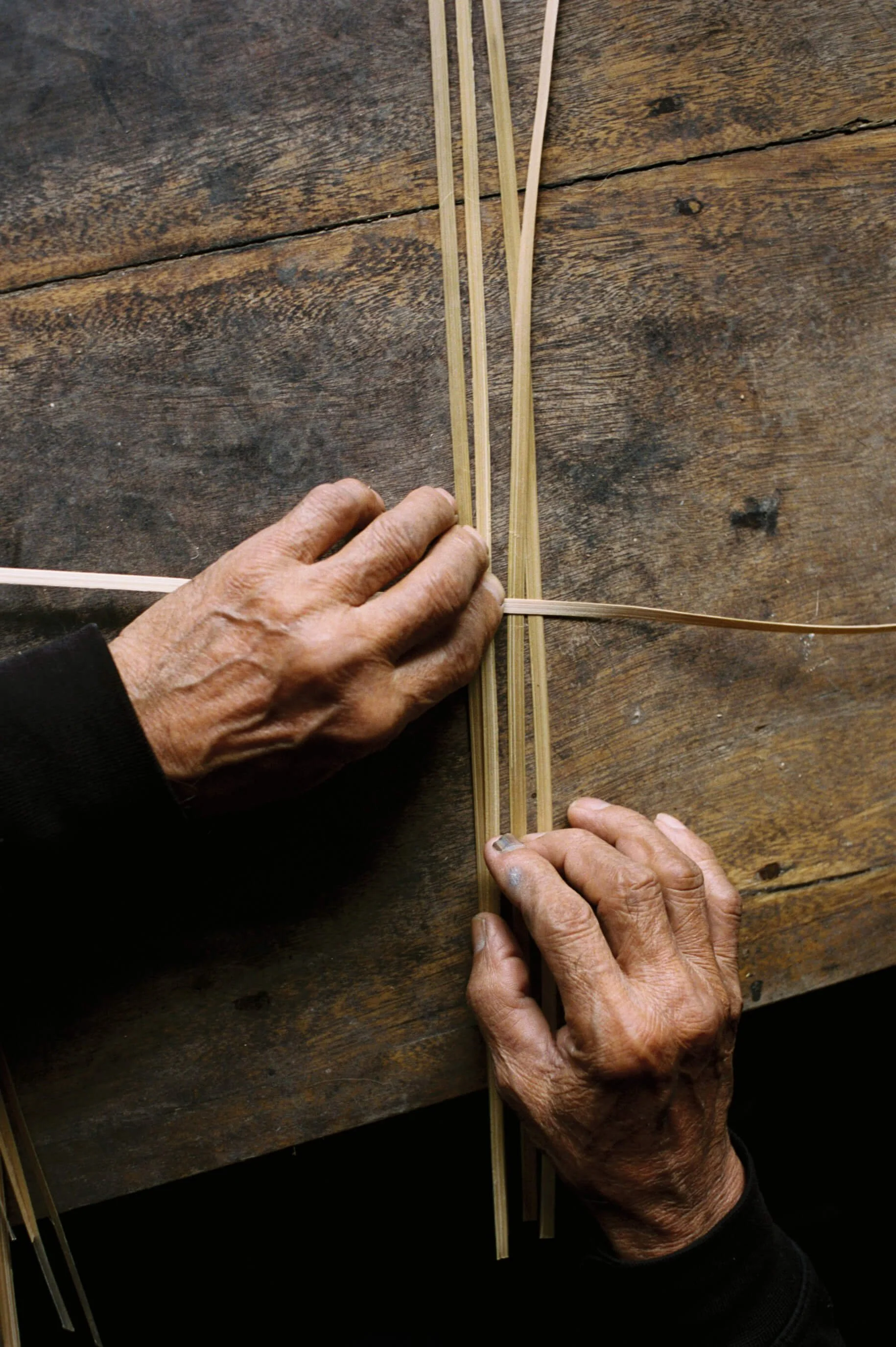 Two elderly hands weaving with thin bamboo strips on a rustic wooden table.