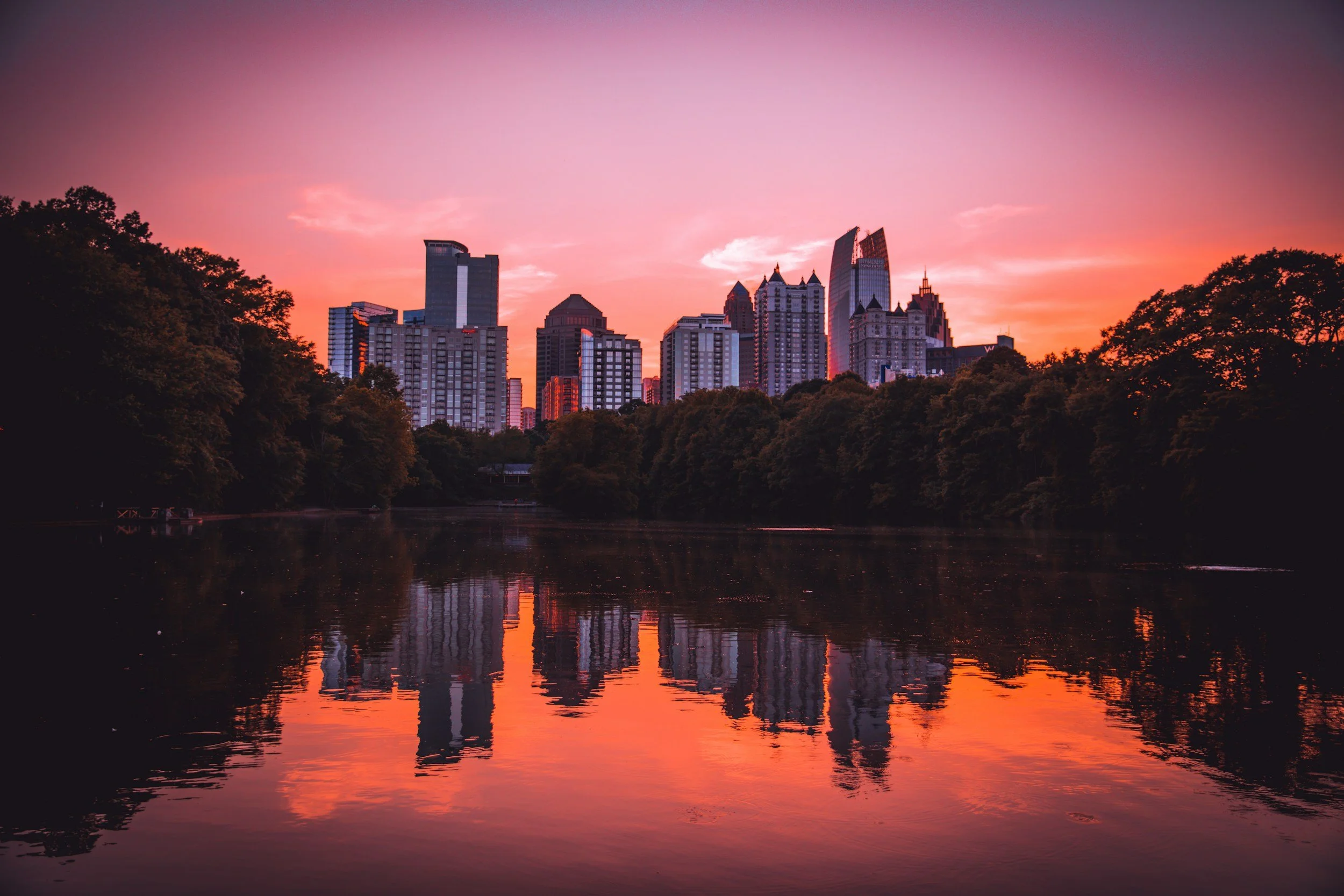City skyline at sunset reflected in a river, with trees on both sides.