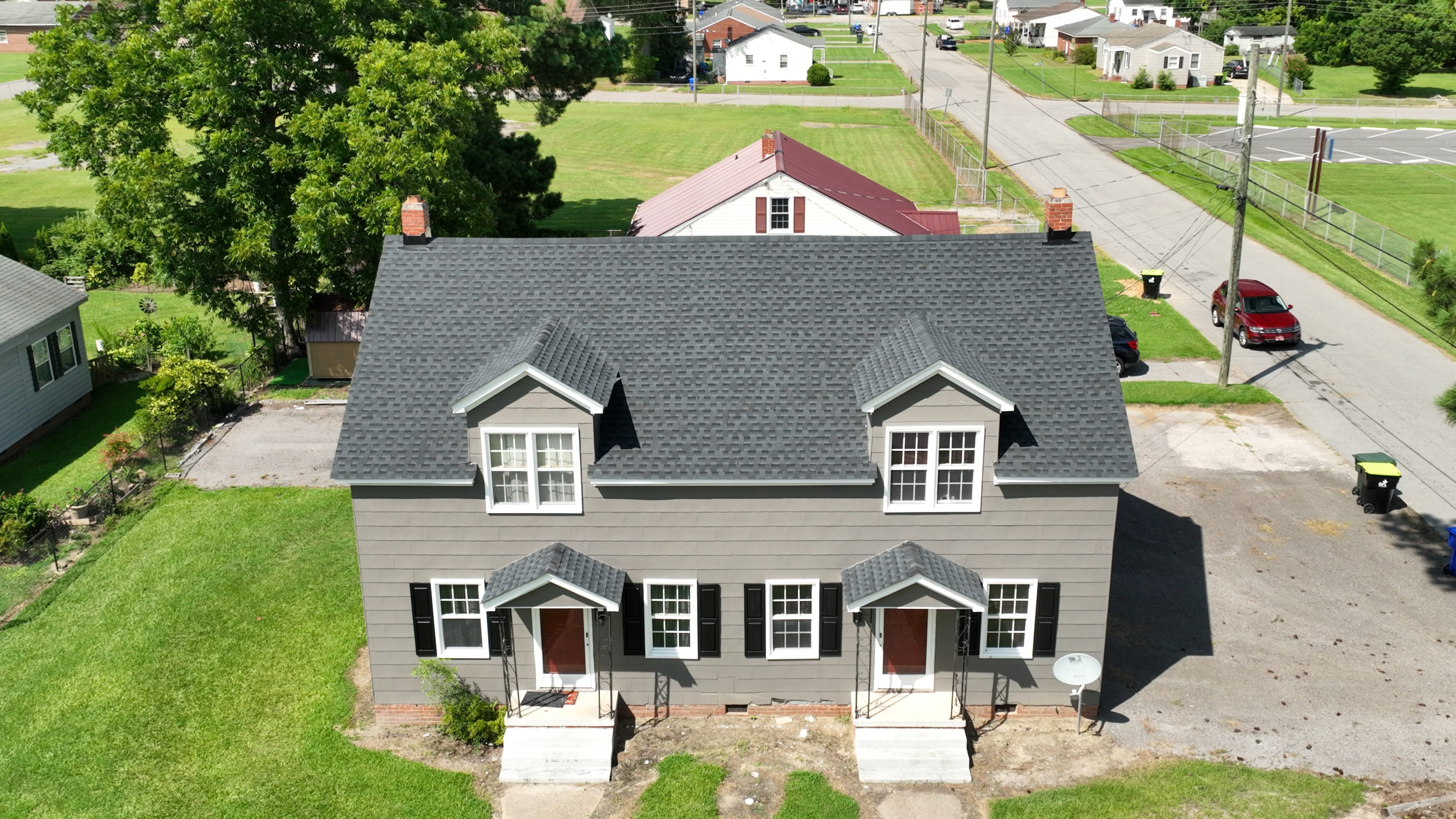 A two-story gray house with black shutters and white trim, featuring two dormer windows and two front porches with steps, situated on a grassy lawn. Surrounding the house are trees, neighboring houses, a parking lot, and a street with cars and trash bins.