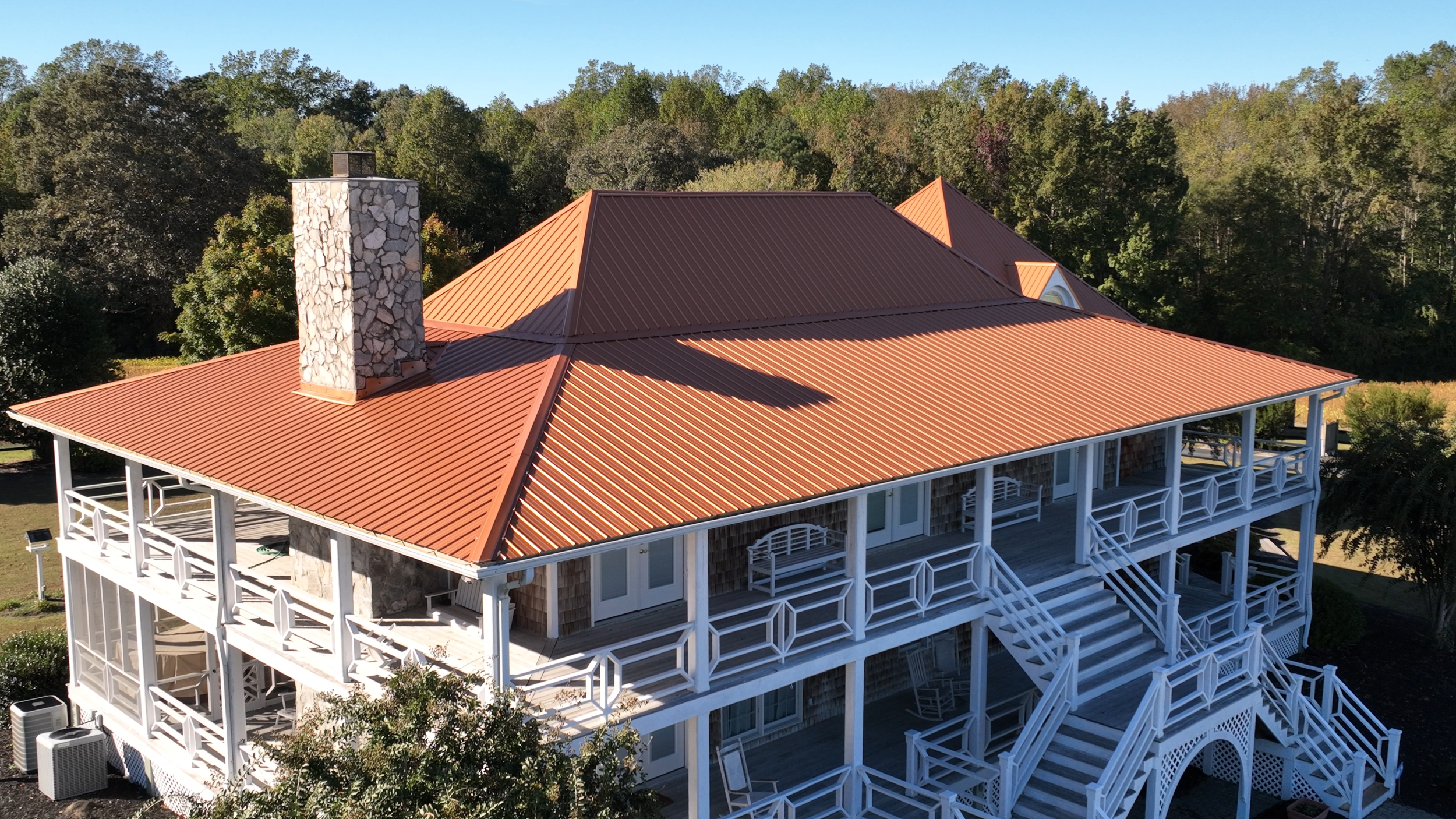 A large house with a red metal roof, white porch railings, and a stone chimney, surrounded by trees.