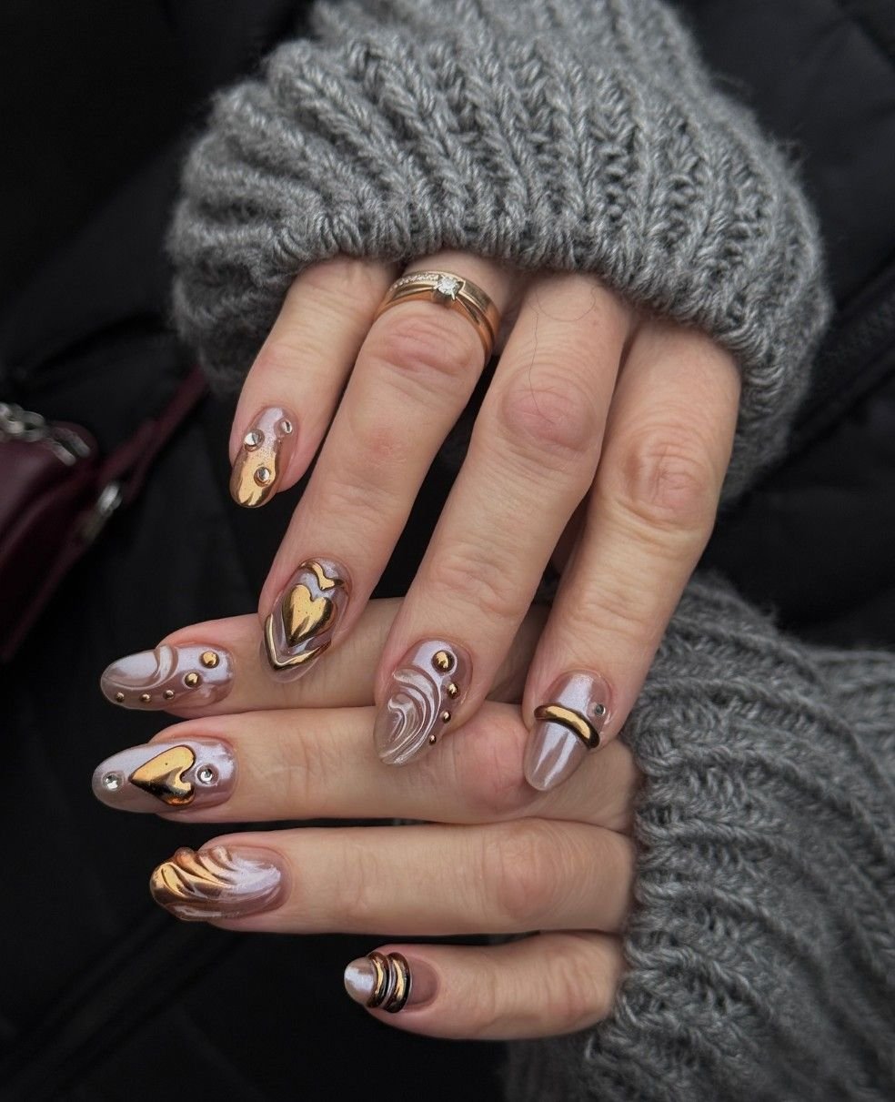 Close-up of hands with elaborate nail art featuring gold, pearl, and marbled designs, with rings and gray knitted fingerless gloves.