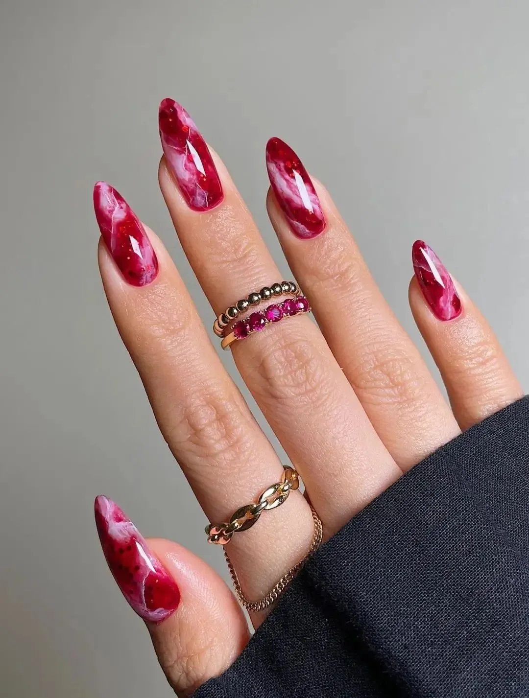 Close-up of a hand with long, marbled red and white nails, wearing multiple rings including gold chains and gemstones, against a plain background.