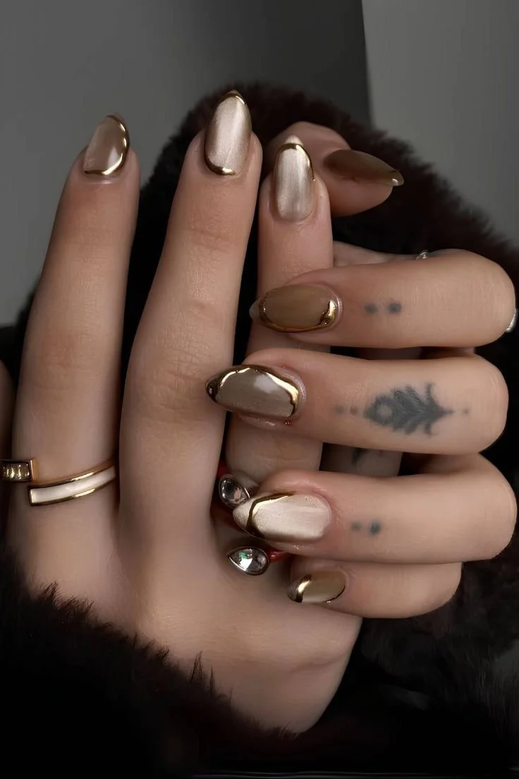 Close-up of a person's hand with metallic gold and beige nail polish, tattoos, and rings, against a dark background.
