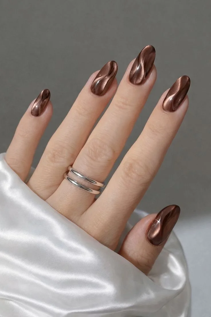 Hand with metallic bronze nail polish and two silver rings on the middle finger, finger partially wrapped in white satin fabric.